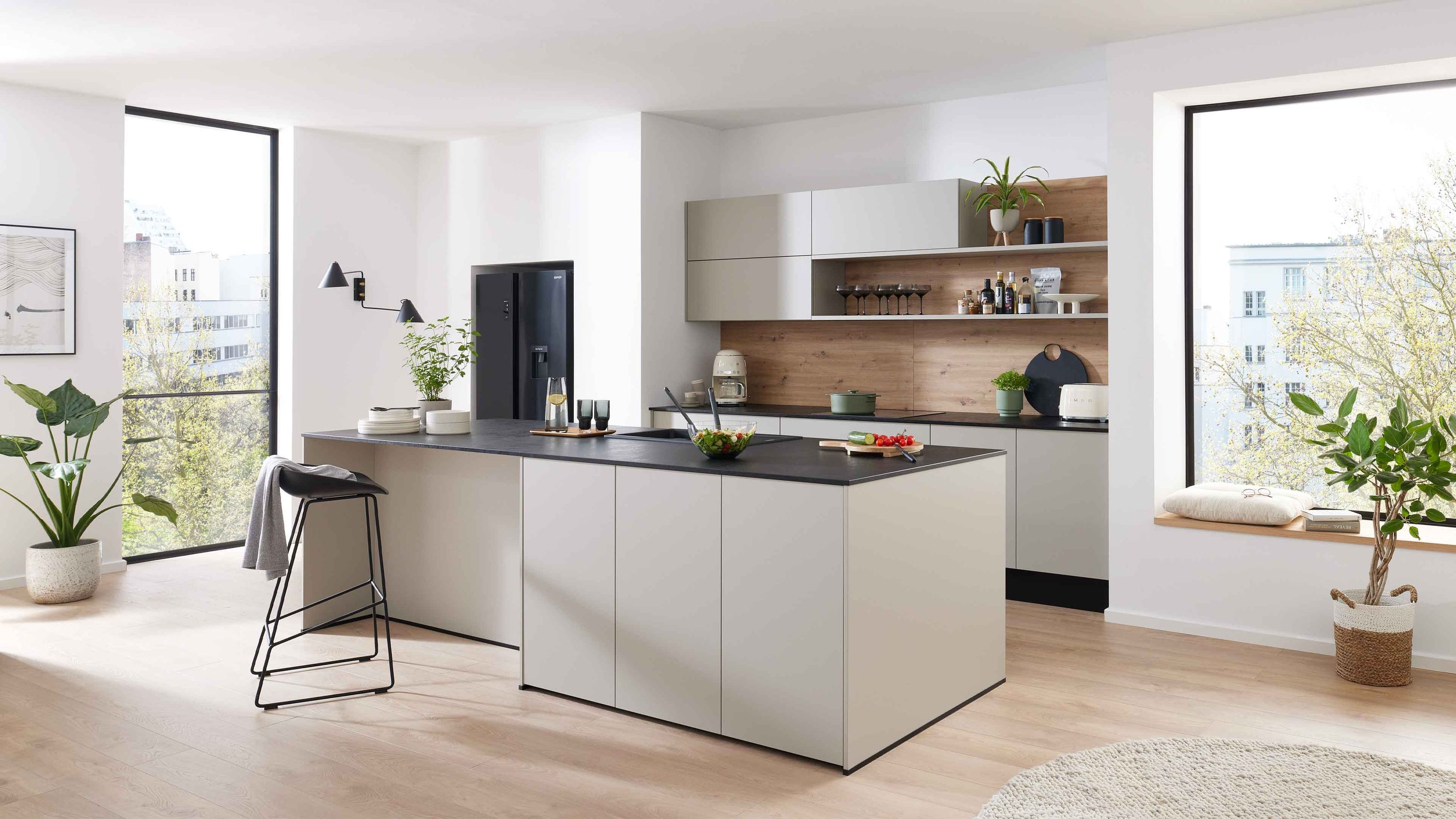 Helle Inselküche in Beige mit schwarzer Arbeitsplatte offenen Holzregalen Einbaukühlschrank und großen Fenstern.