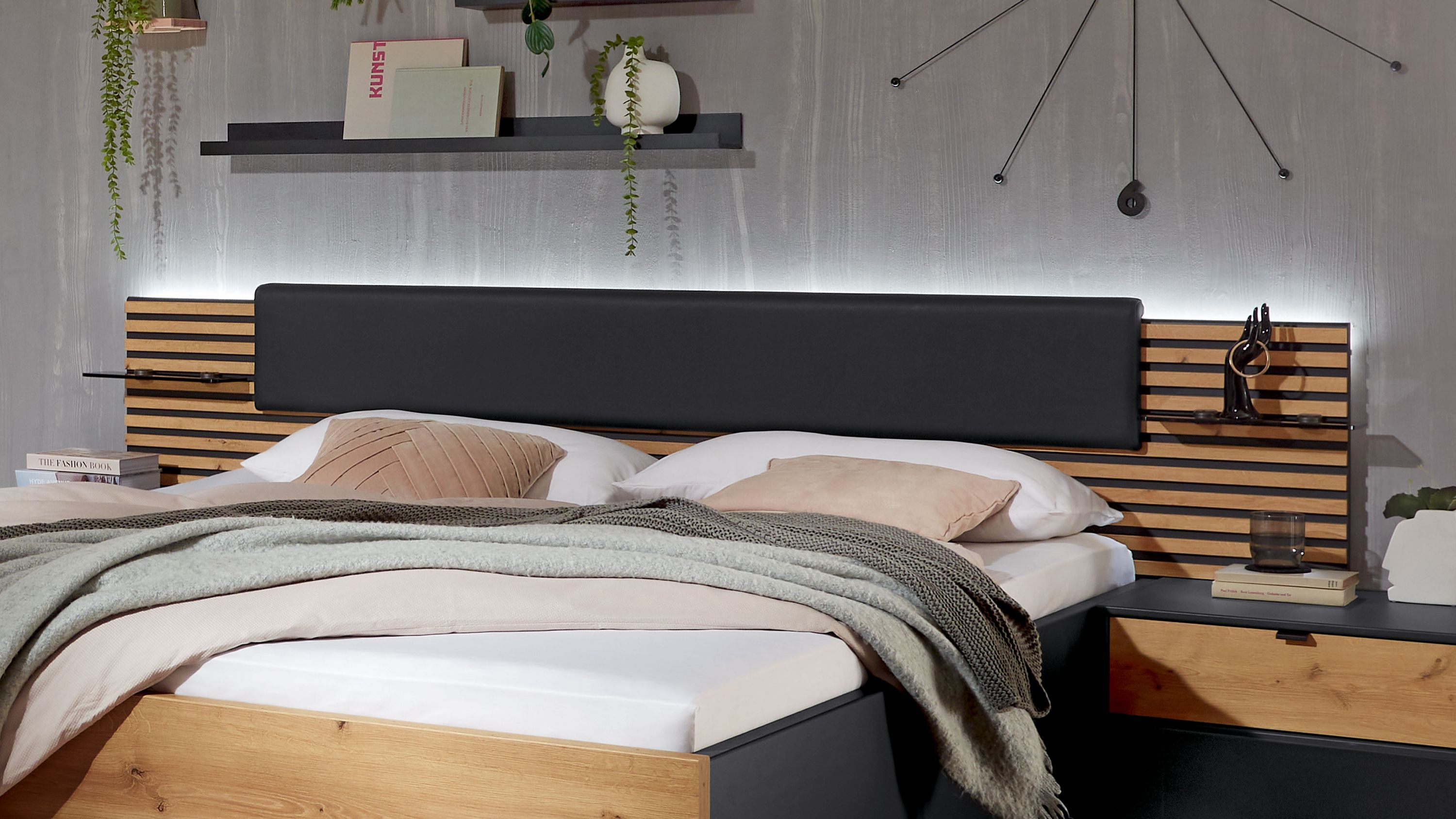 Bett mit beleuchtetem Kopfteil und seitlichen Ablagen Bett mit Holzlatten-Kopfteil und schwarzem Polsterelement seitliche Ablagen mit Schubladen in Holz und dunkler Oberfläche