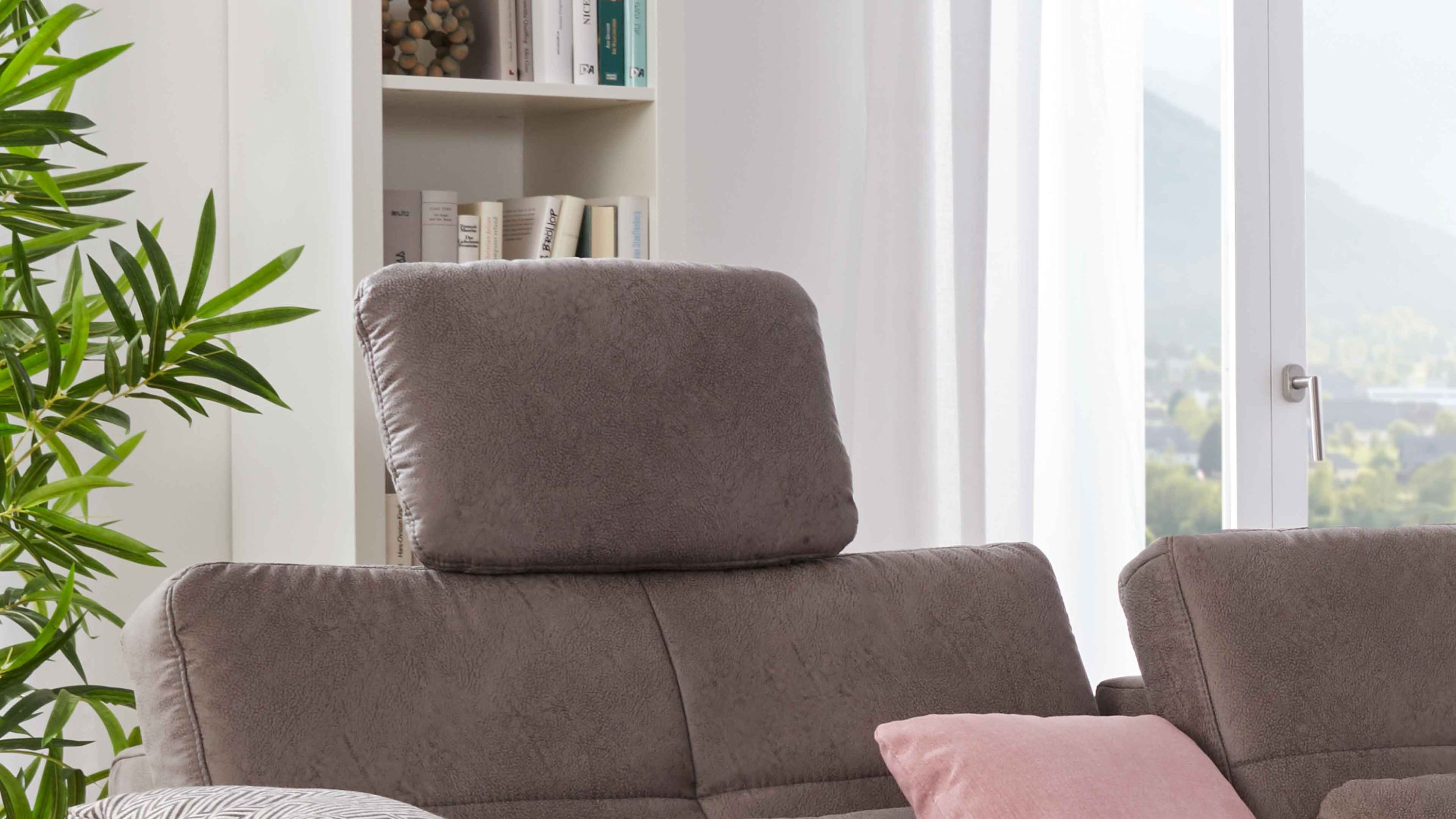 Kopfstütze in grauer Mikrofaser auf einem Sofa in einem hellen Raum mit Fenster und Pflanze