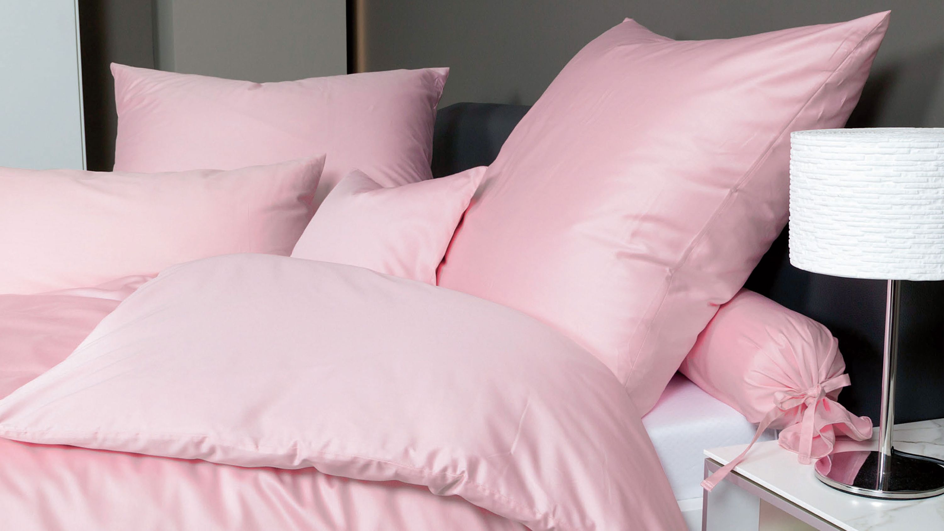 Rosa Bettwäsche-Set mit Kissen und Decke auf einem Bett neben einem Nachttisch mit weißer Lampe
