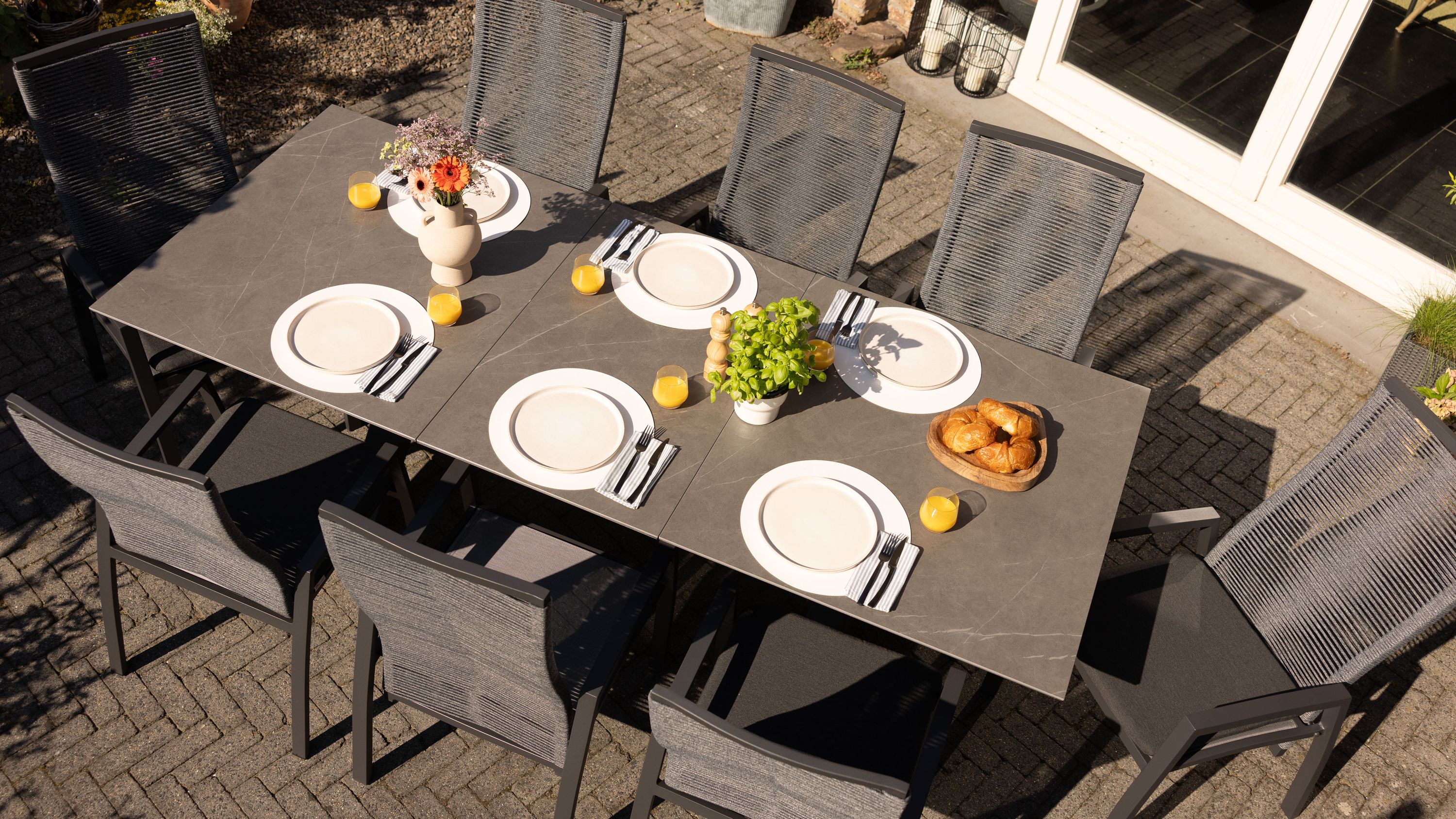 Rechteckiger Ausziehtisch mit grauer Tischplatte und anthrazitfarbenen Stühlen auf gepflasterter Terrasse