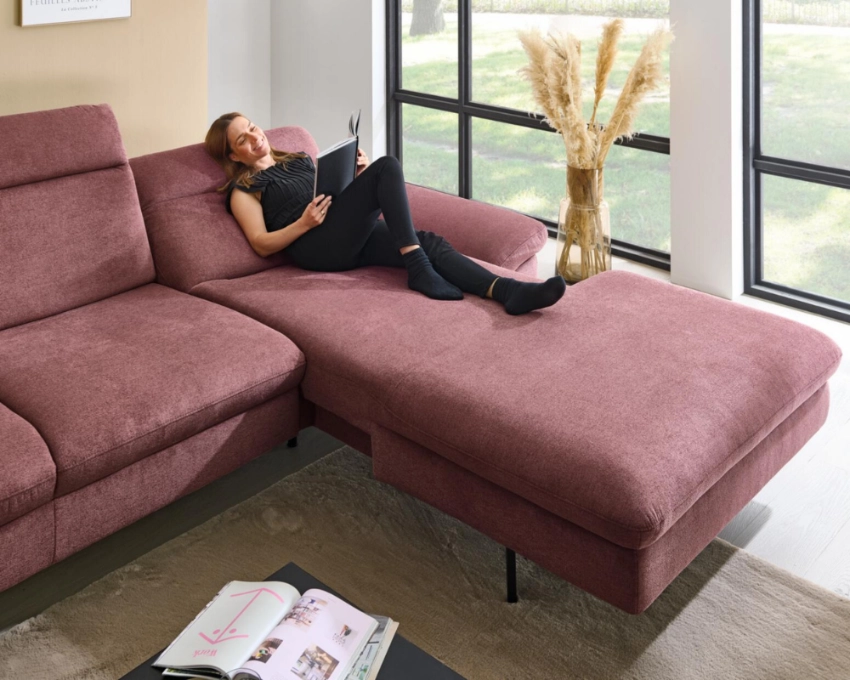 Entspanntes Arbeiten im Wohnzimmer auf modernem Ecksofa mit Laptop und Tageslicht-Atmosphäre Frau liegt entspannt auf rosa Ecksofa mit Laptop, große Fenster im Hintergrund, modernes Wohnzimmer-Design.