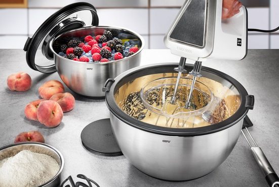 Küchenmixer rührt Teig, daneben Schüsseln mit Beeren und Mehl, Pfirsiche liegen auf der Arbeitsplatte, frontal aufgenommen.