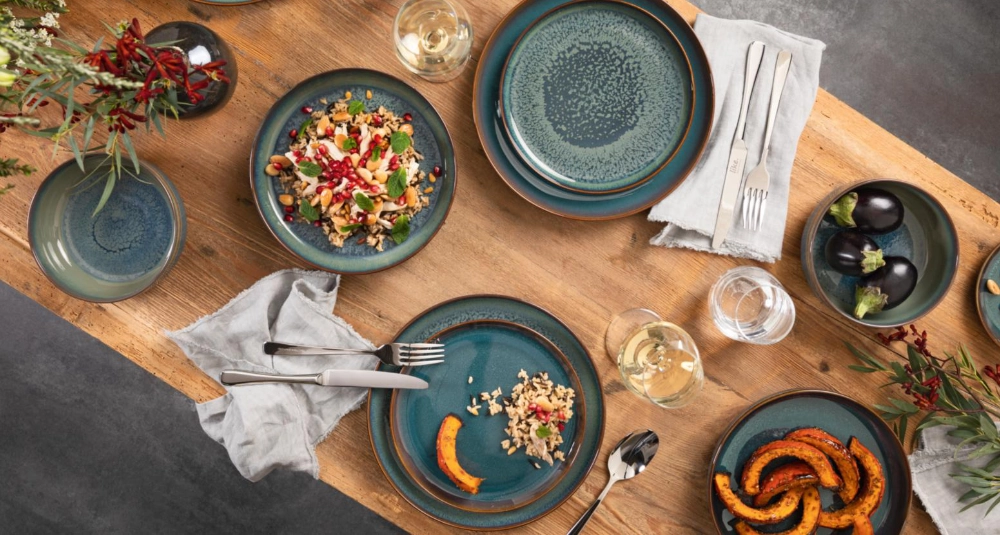 Gedeckter Holztisch mit blau-grünem Steingutgeschirr, herbstlichen Speisen, Servietten, Weingläsern und Blumendekoration.