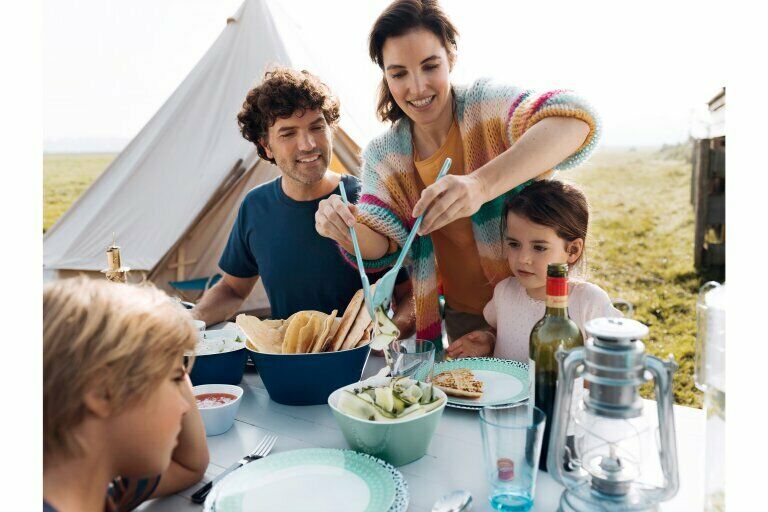 Gruppe isst draußen neben Zelt, Tisch mit Brot, Salat und Getränken gedeckt, sommerliche Atmosphäre