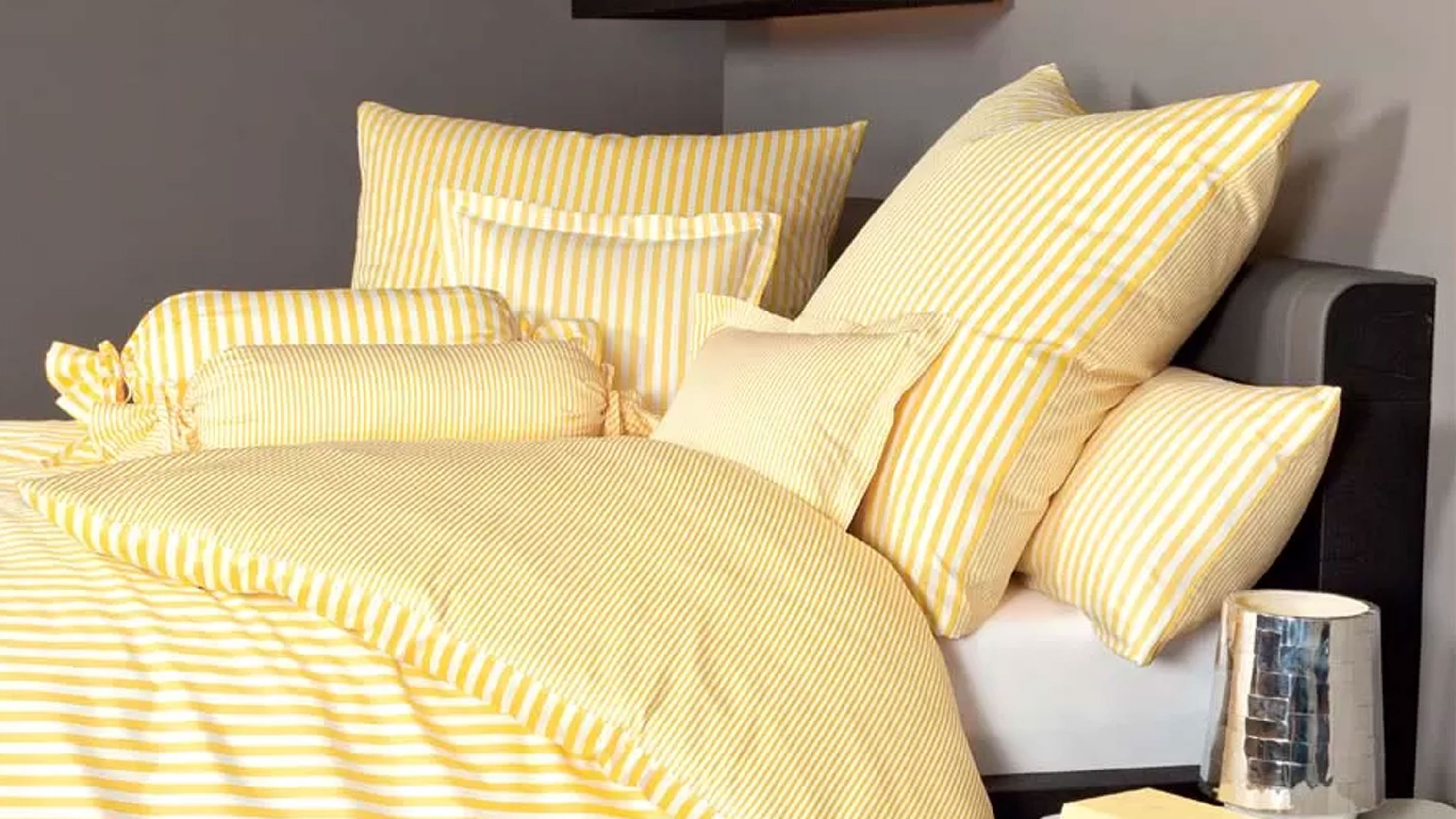 Hellgelbes Bettwäsche-Set mit Streifenmuster auf Bett Hellgelbe Bettwäsche mit verschiedenen Kissen in Streifenmuster auf einem Bett mit dunklem Kopfteil