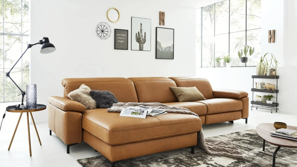 Modernes Wohnzimmer mit großem cognacfarbenem Ledersofa, dekoriert mit Kissen und Decken. Helle, luftige Einrichtung mit Wandbildern, Pflanzen und Teppich im skandinavischen Stil für gemütliches Wohnen.