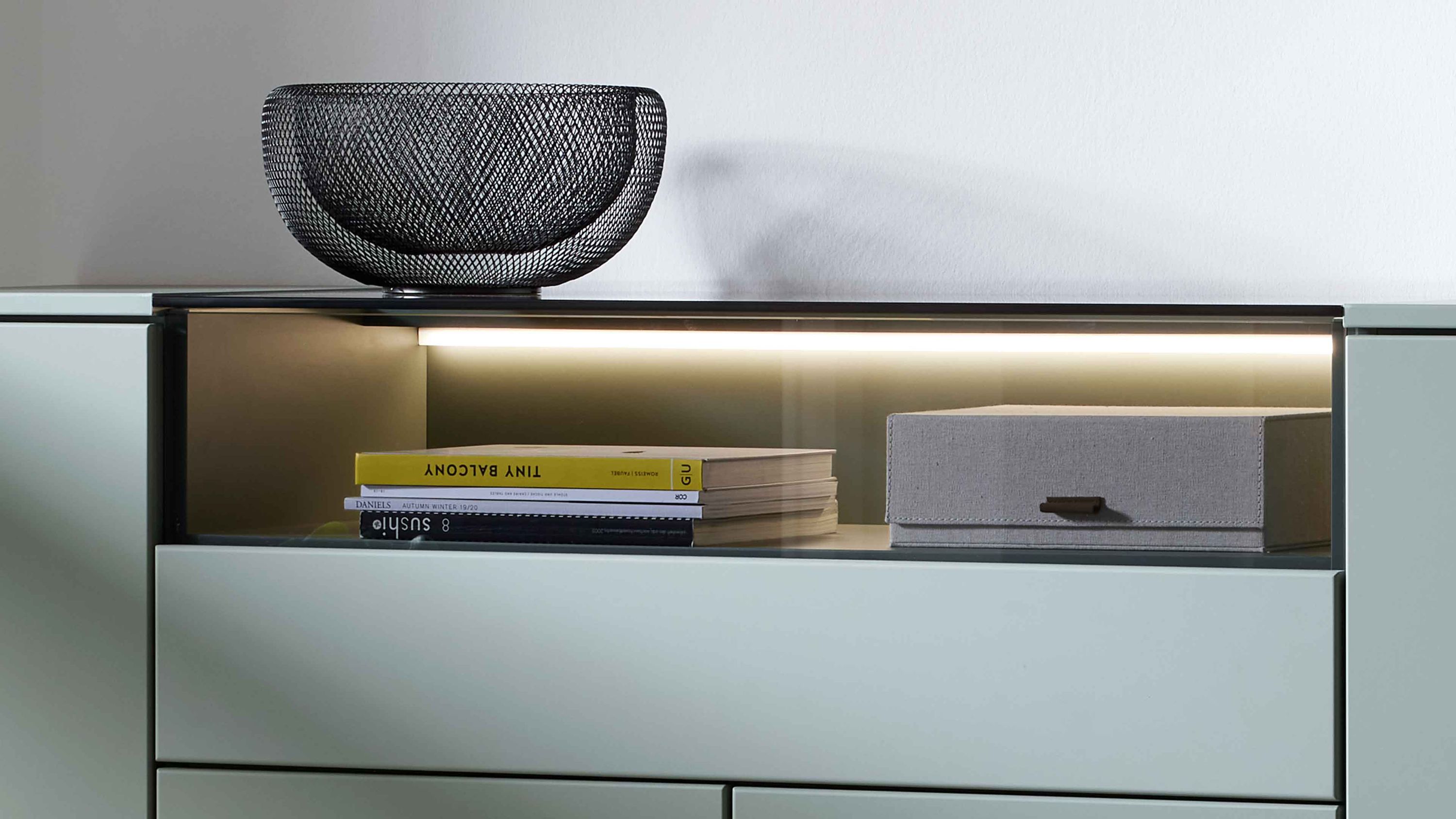 Wohnzimmerschrank mit LED-Beleuchtung offenem Fach mit Büchern und grauer Box schwarze Drahtschale auf der Ablage