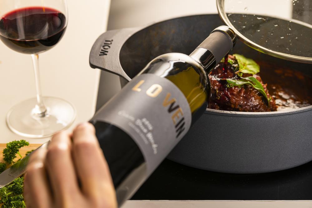 Anthrazitfarbener Gussbräter Hand gießt Rotwein hinein Glas daneben leicht schräg von oben rechts aufgenommen