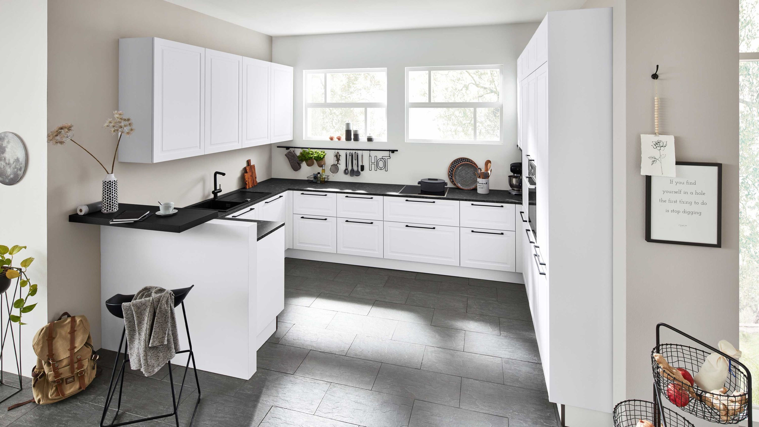 U-Küche mit Kontrastflächen und natürlichem Lichteinfall U-Küche mit weißen Schränken schwarzen Arbeitsflächen Fensterlicht Hocker und Wandbild neben Kühlschrank