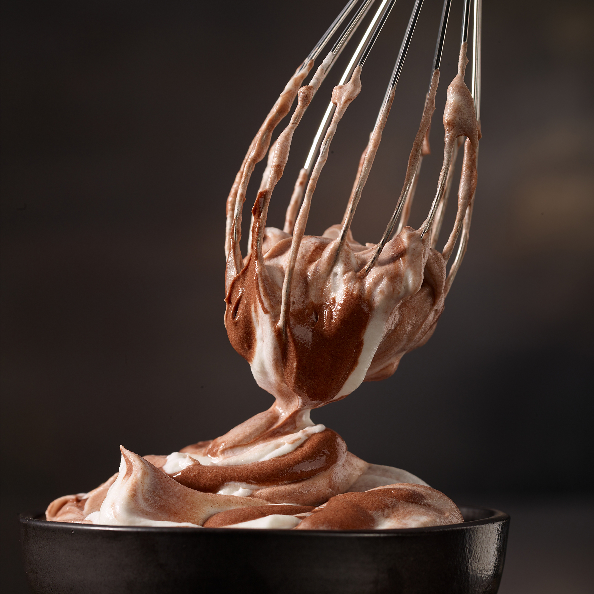 Schneebesen silberfarben mit Schokocreme über Schüssel gehalten weitere Creme darin dunkler Hintergrund schräg vorn