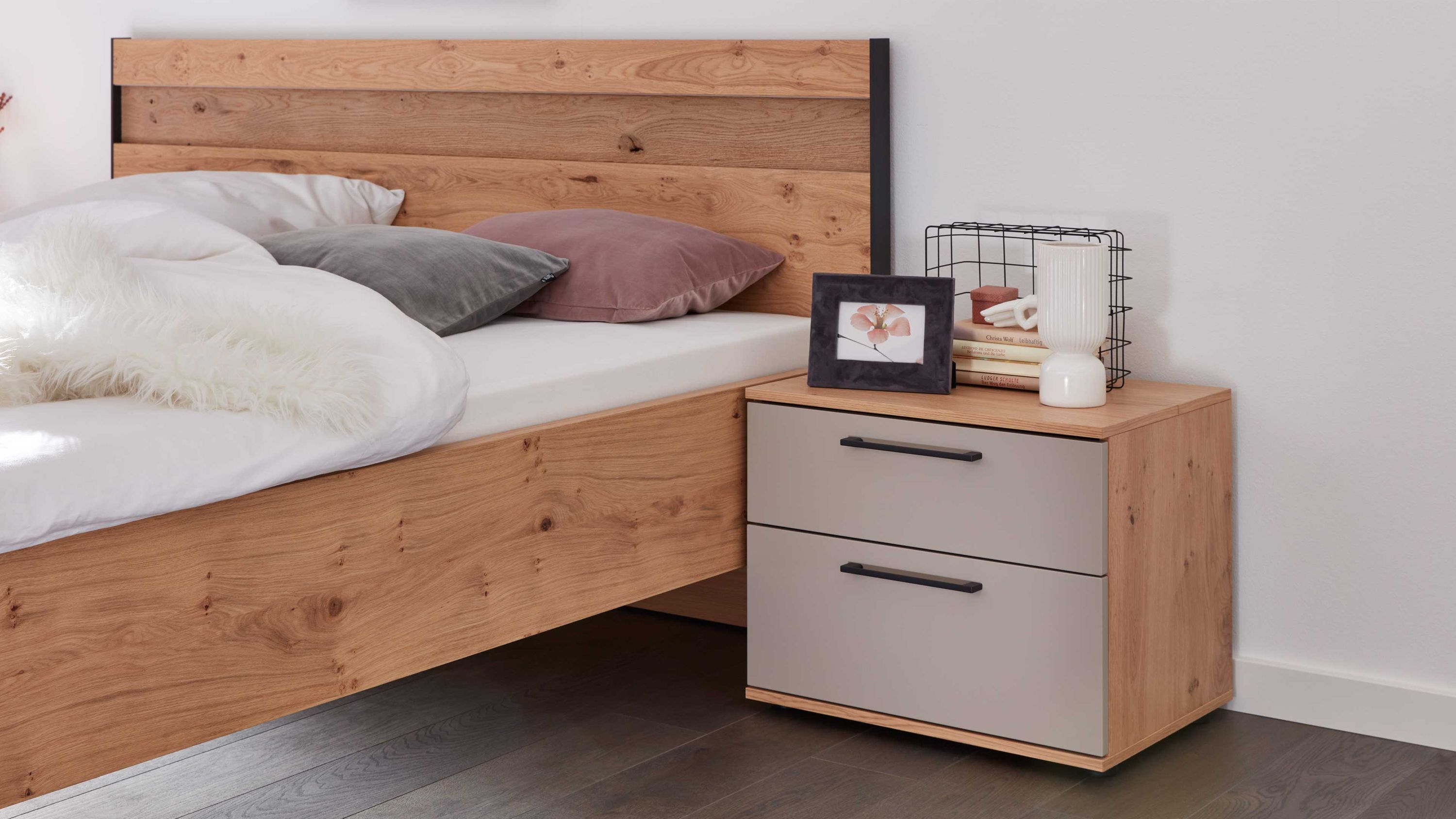 Nachtkommode mit zwei taupefarbenen Schubladen und Holzoptik seitlich neben einem Bett mit Holzkopfteil