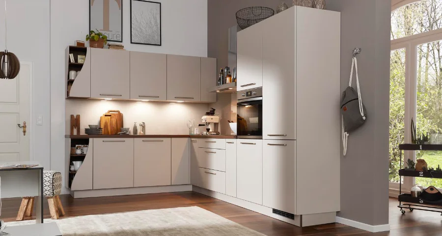 Moderne L-förmige Küche in warmen Beigetönen mit Hängeschränken, offenen Regalen und Einbaugeräten, gemütlich beleuchtet in einem hellen Raum mit Holzboden und großen Fenstern.