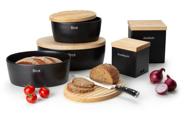 Schwarze Vorratsdosen mit Eichenholzdeckeln beschriftet umgeben von Brot Tomaten und Zwiebeln