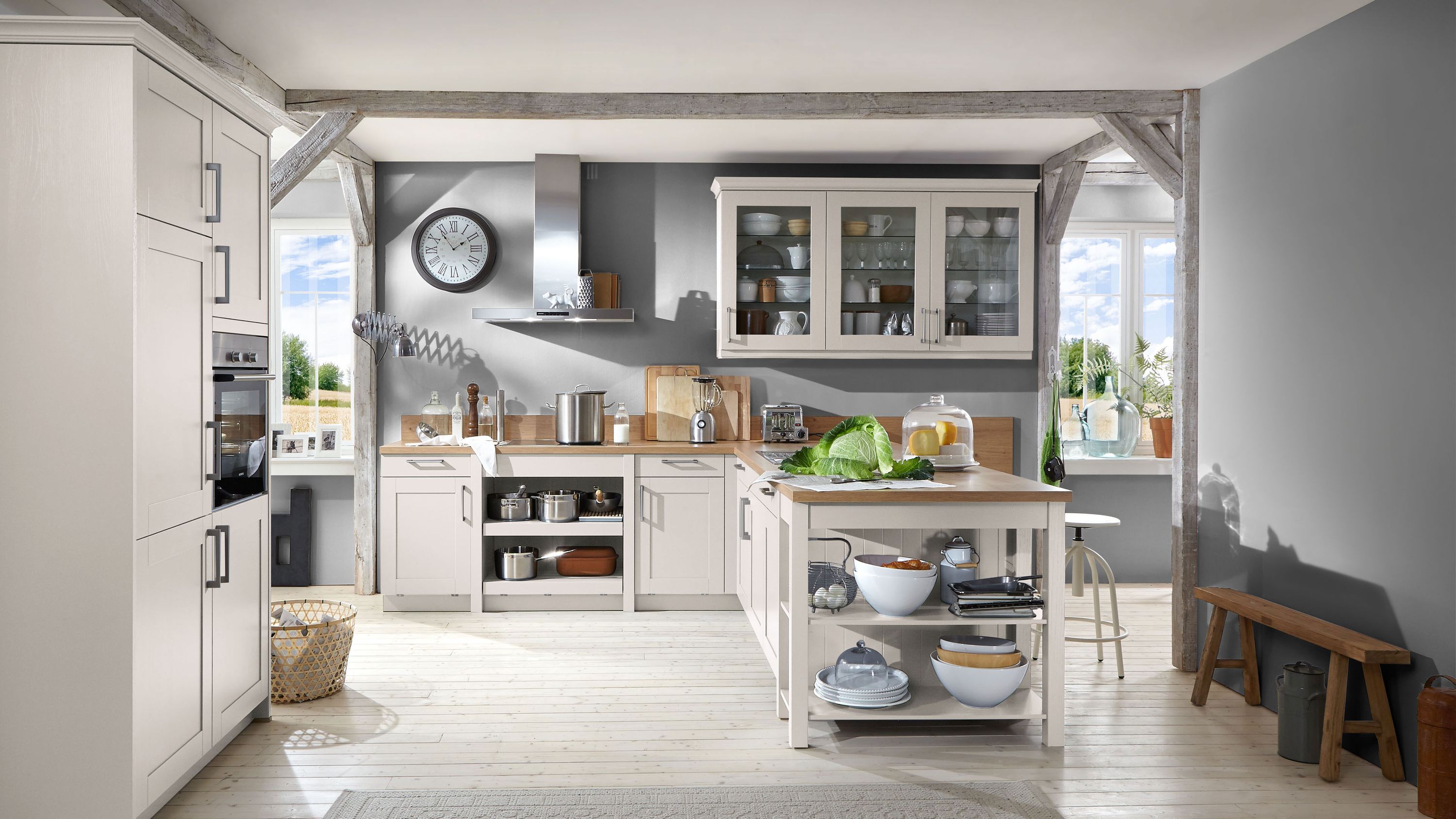 Weiße Landhausküche mit Kochinsel und Glashängeschränken. Weiße Landhausküche mit Kochinsel offenen Regalfächern Glashängeschränken und Holzbalken im Raum.