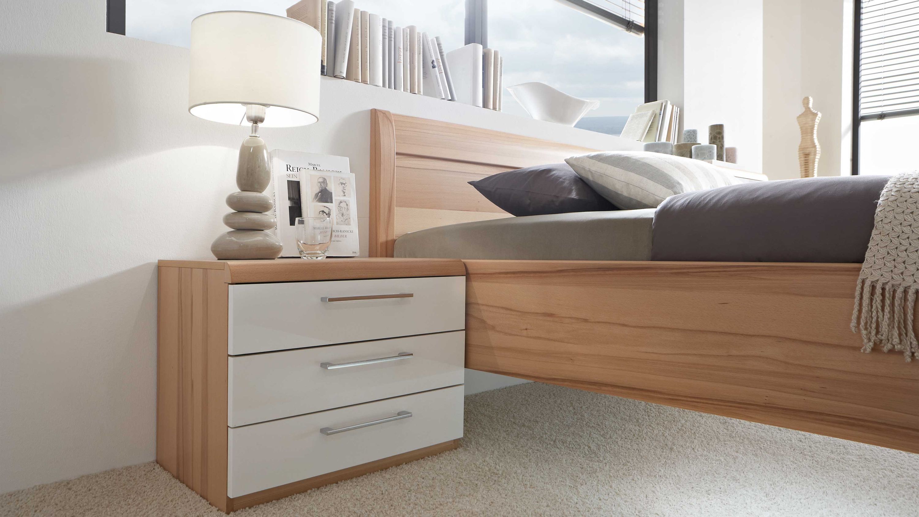 Nachtkommode mit drei Schubladen in Weiß und hellem Holz Nachtkommode mit drei weißen Schubladen und hellem Holzrahmen neben einem Bett mit Kopfteil aus hellem Holz