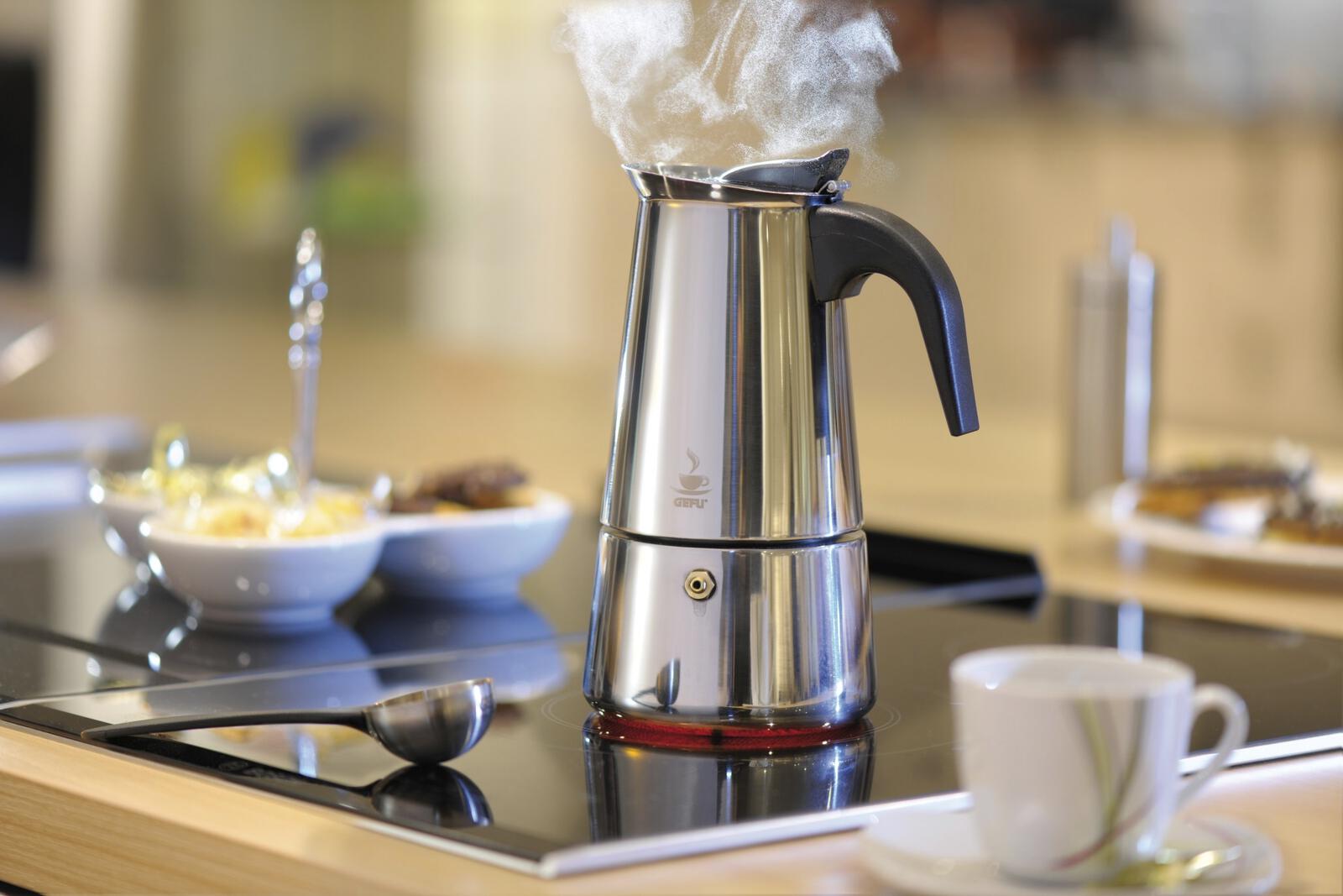 Aus leicht schräger Perspektive: Edelstahl-Kaffeekocher auf Arbeitsplatte, dahinter Tasse, Untertasse und zwei Schalen
