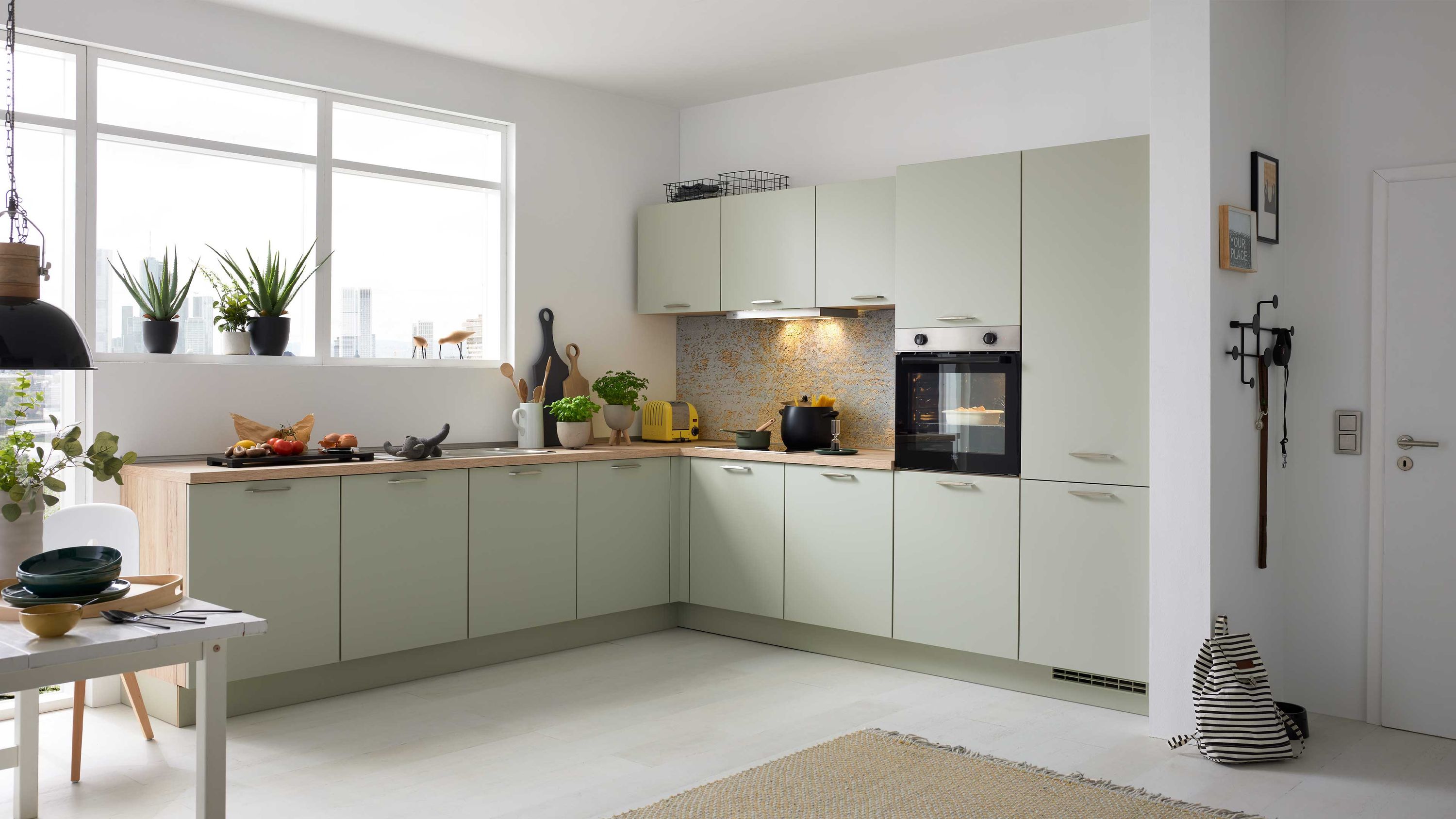 Helle L-Küche mit Pflanzen und Frühstückstisch Helle Fronten L-Form Fenster mit Pflanzen Einbauofen; Teppich Tisch mit Geschirr links Küchenutensilien auf Arbeitsfläche
