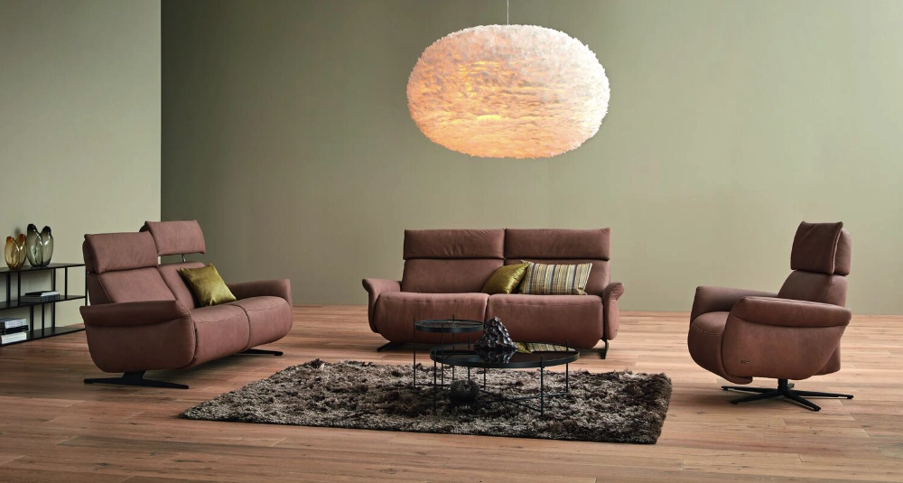 Modernes Wohnzimmer mit braunem Ledersofa, zwei Relaxsesseln, flauschigem Teppich und großer Deckenleuchte.