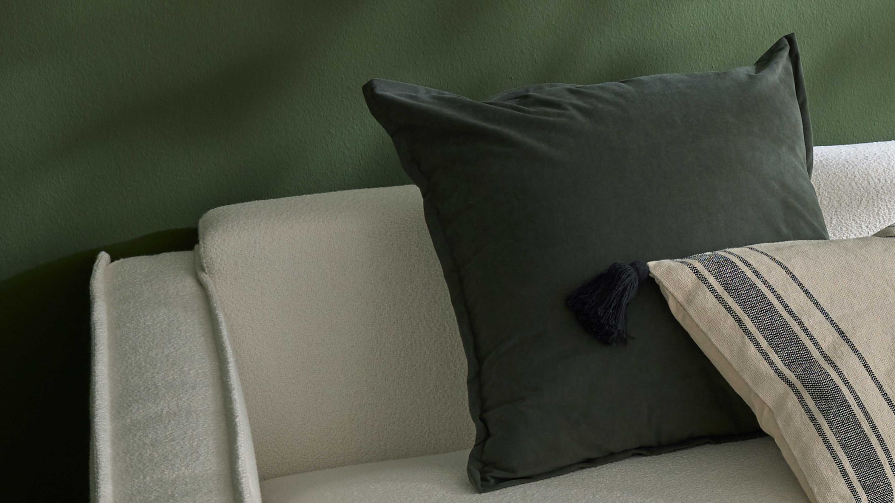 Grünes quadratisches Kissen auf Sofa Quadratisches Kissen in dunklem Grün auf hellem Sofa vor grüner Wand