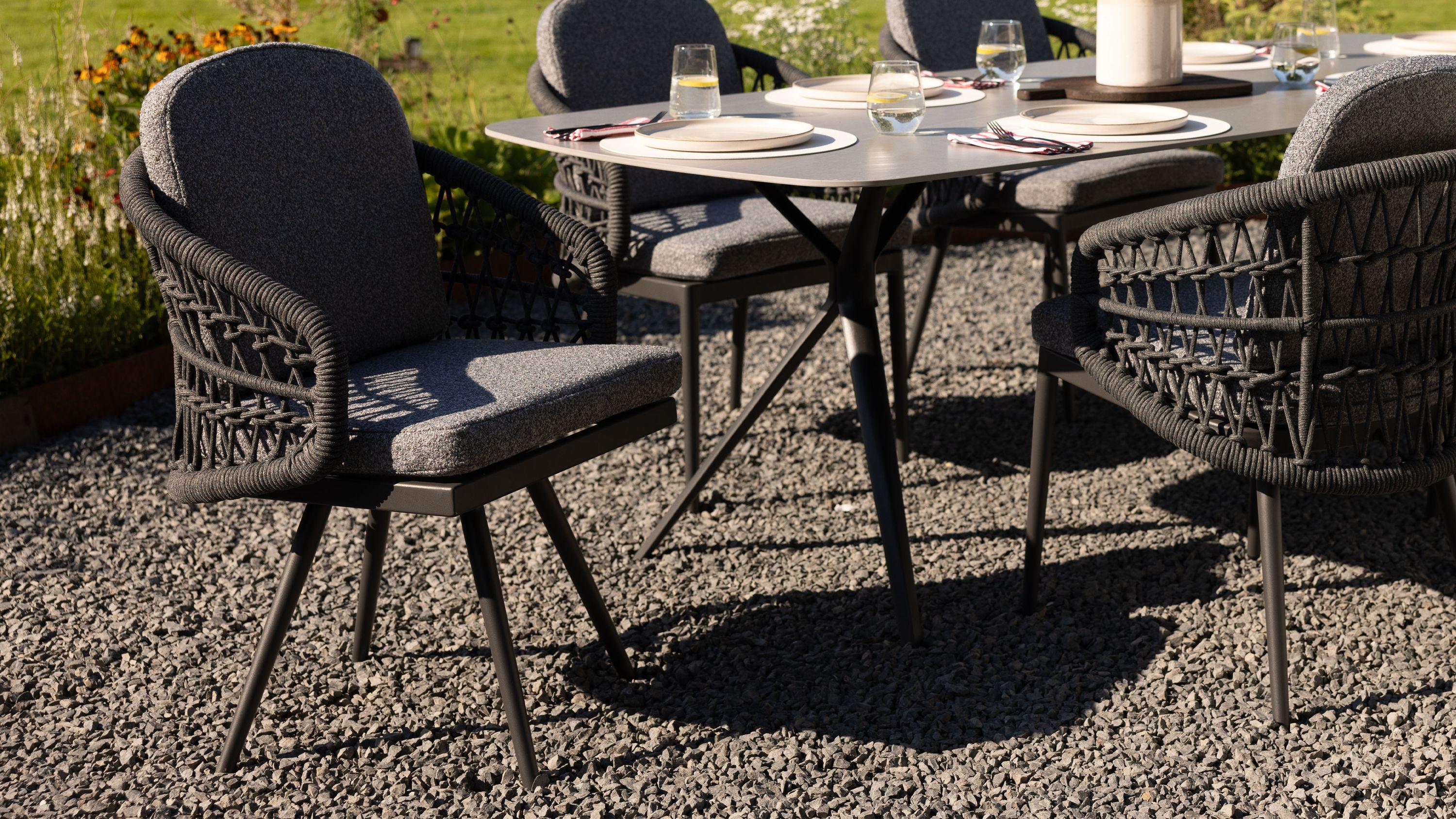 Drehsessel mit grauem Polster und anthrazitfarbenem Geflecht an einem Gartentisch auf Kiesboden