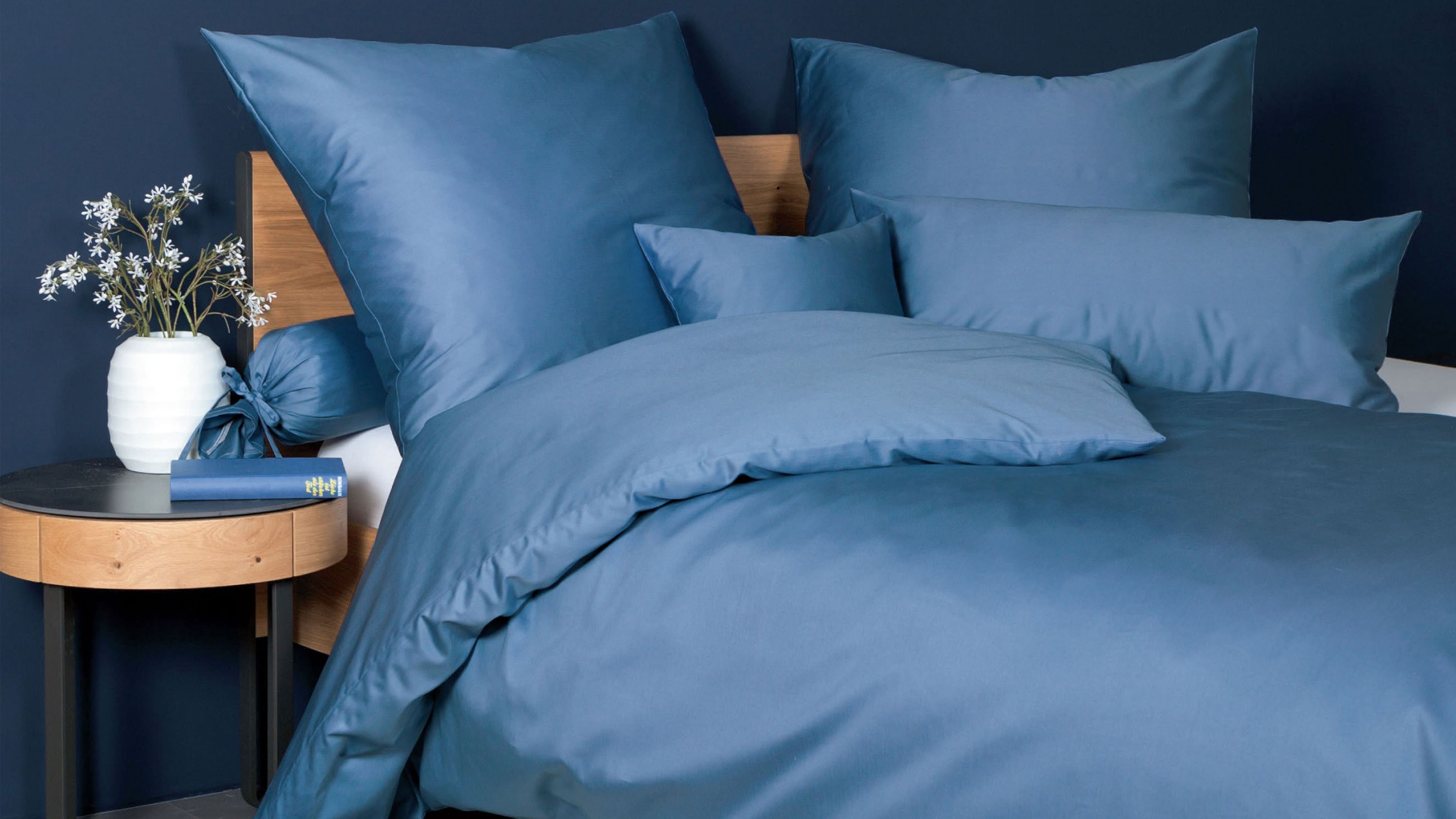 Bett mit einfarbiger blauer Bettwäsche und mehreren Kissen vor dunkelblauer Wand