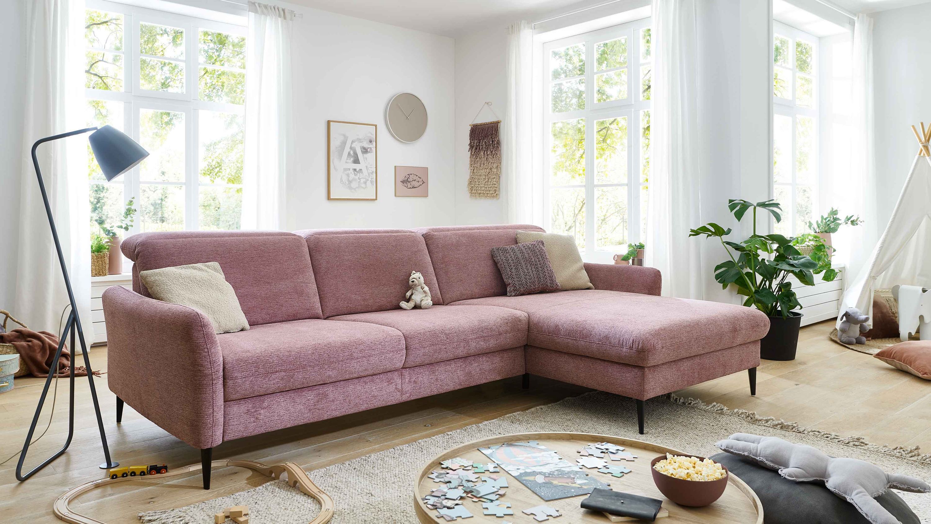 Ecksofa mit rosafarbenem Stoffbezug und schwarzen Füßen in hellem Wohnzimmer mit Kissen und Teppich