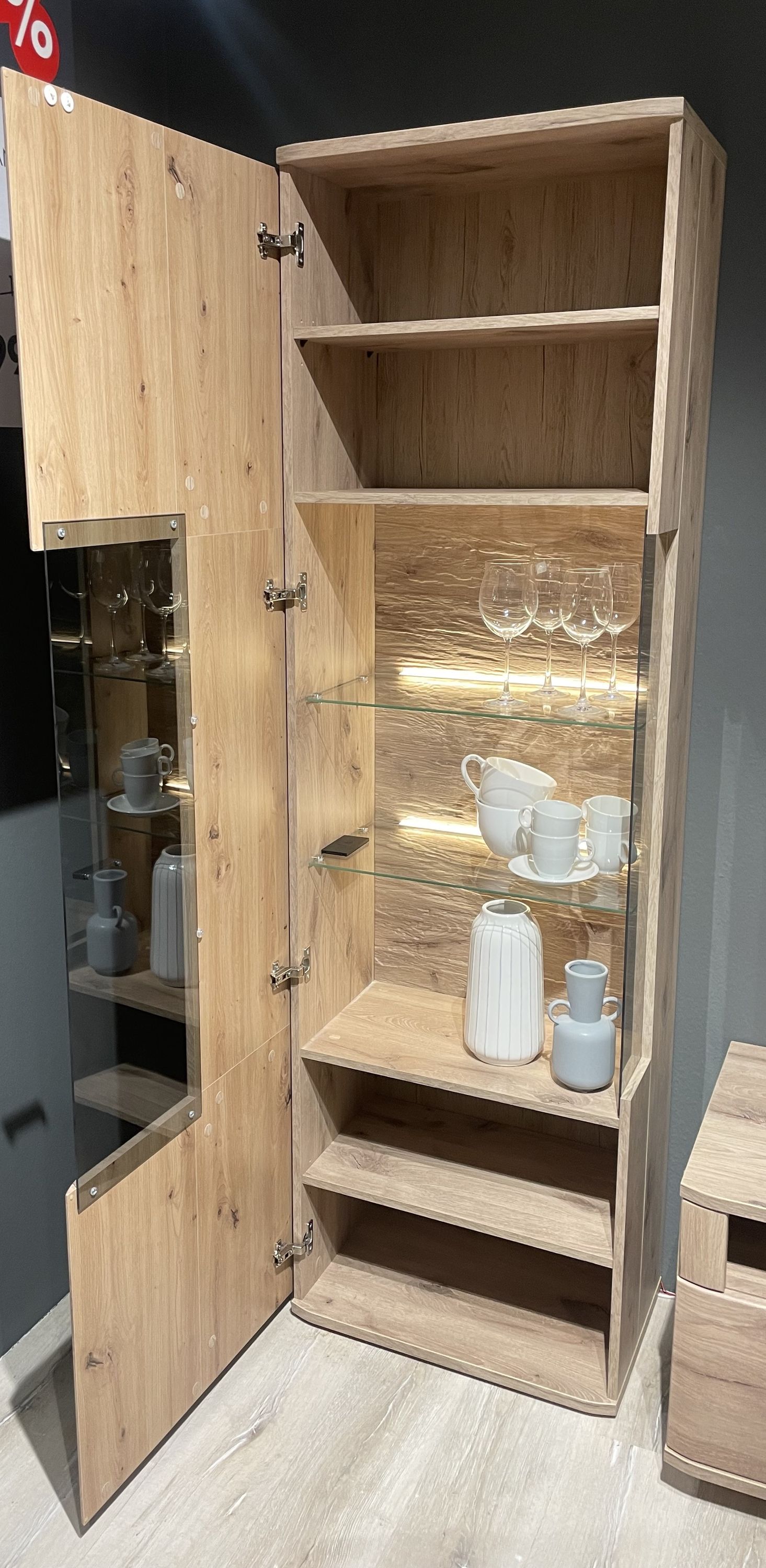 Hoher Schrank mit offenem Türfach Holzoptik zwei Glasböden mit Gläsern und Geschirr darunter offene Regalfächer