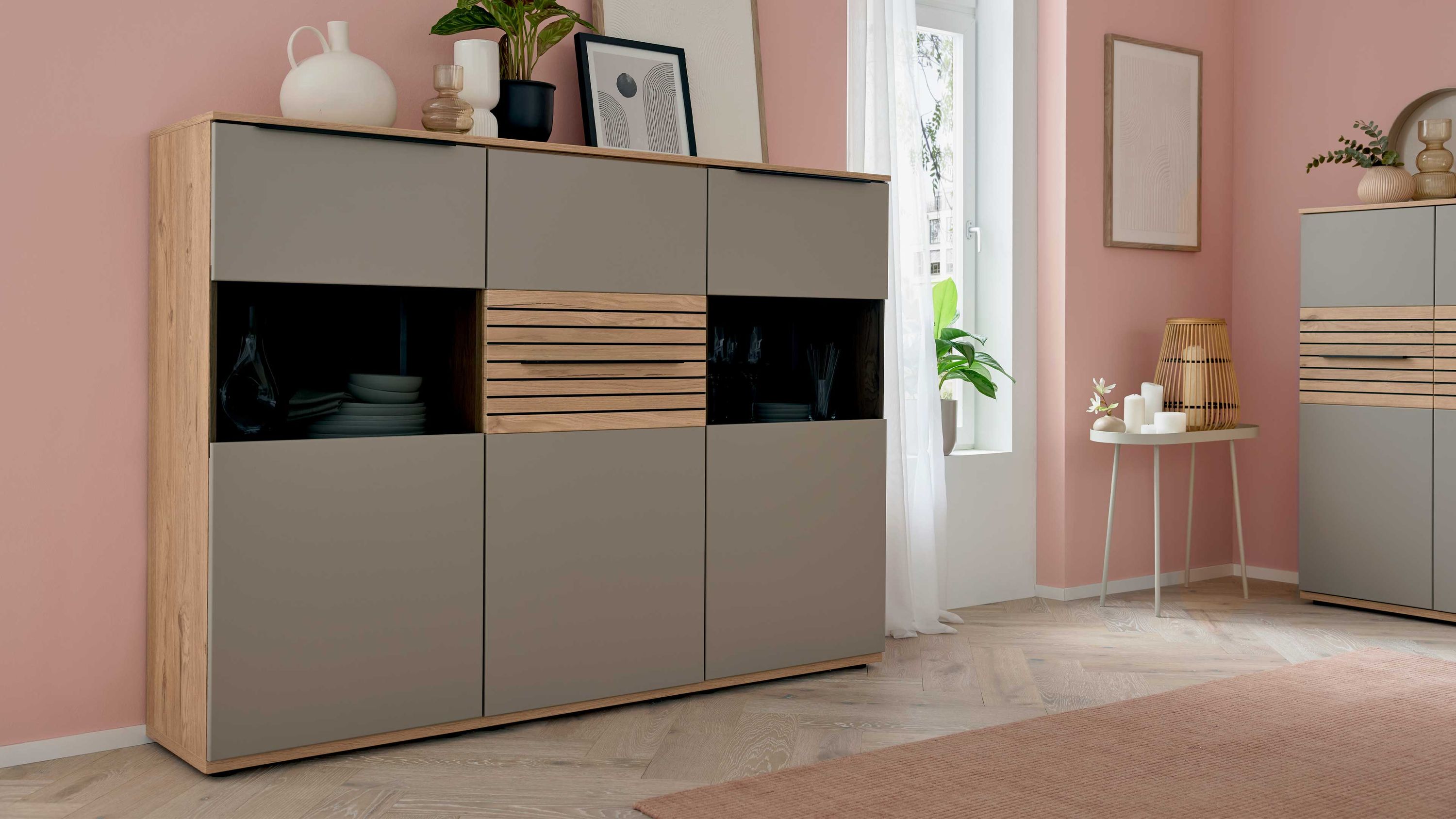 Highboard mit drei grauen Türen und mittleren Fächern mit Holzlamellen und Glasfront in einem Wohnzimmer mit rosa Wänden