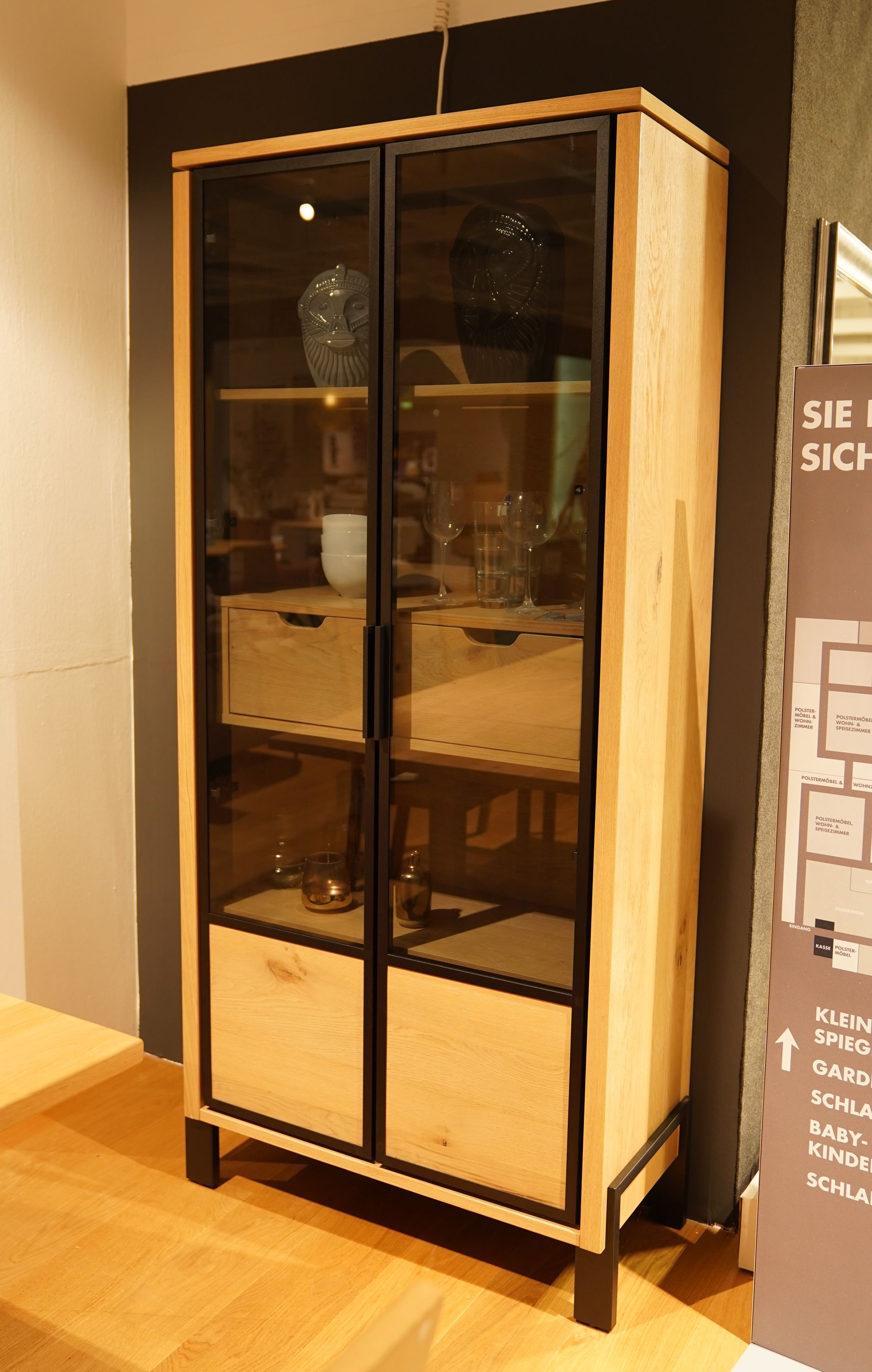 Hölzerne Vitrine mit Glastüren, zwei Schubladen und schwarzem Metallrahmen, dekoriert mit Gläsern und Keramik.