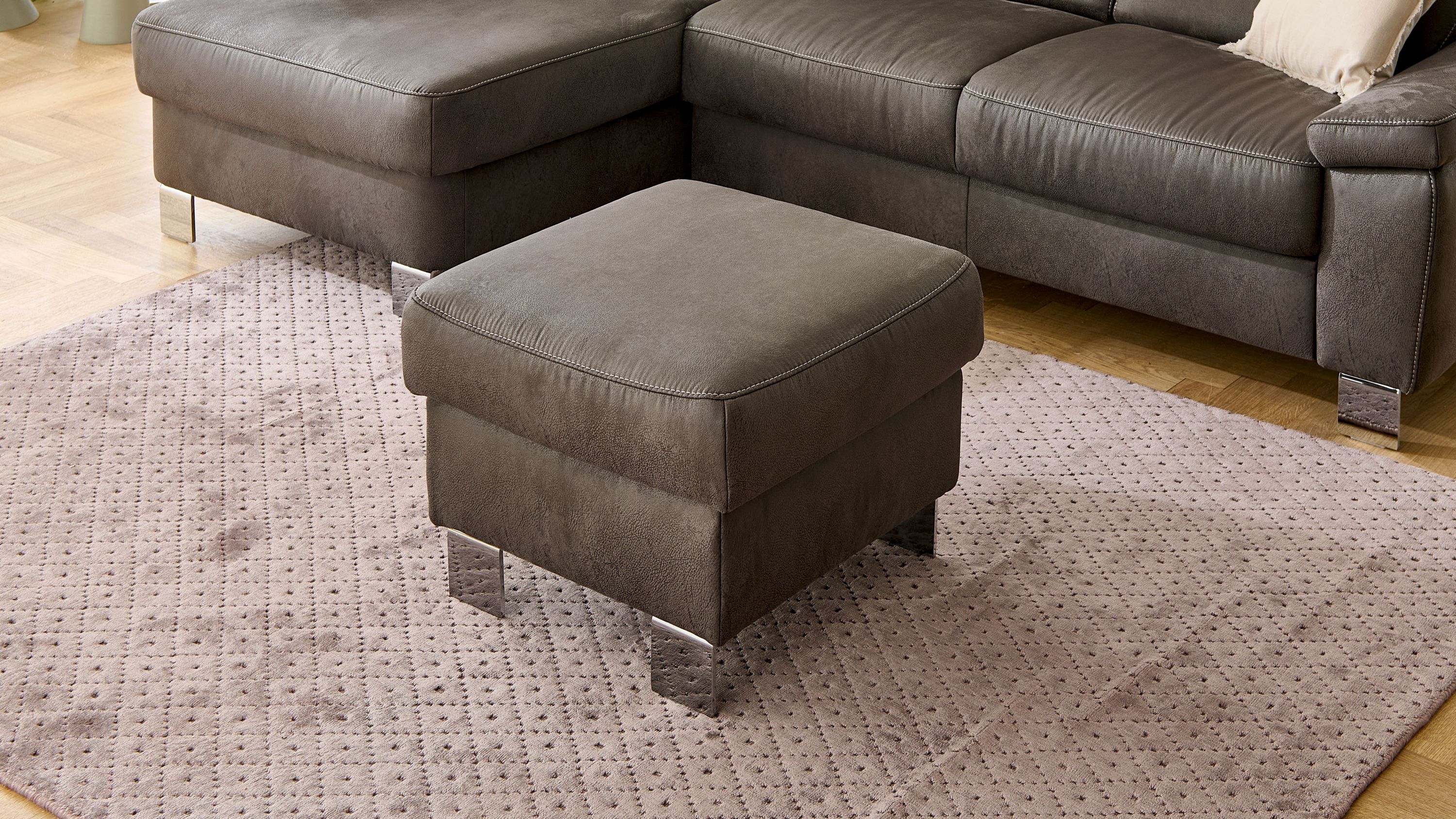Quadratischer Polsterhocker in anthrazit mit Metallfüßen auf gemustertem Teppich vor grauem Sofa