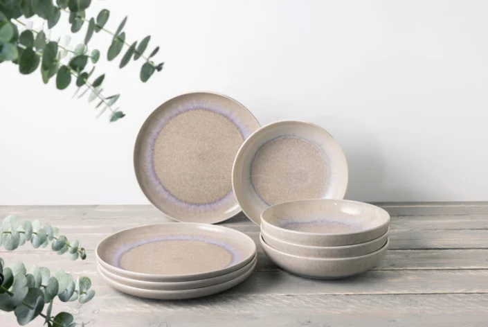 Beigefarbenes Steingut-Geschirrset mit dezentem Randmuster, bestehend aus Tellern und Schalen auf Holzoberfläche.