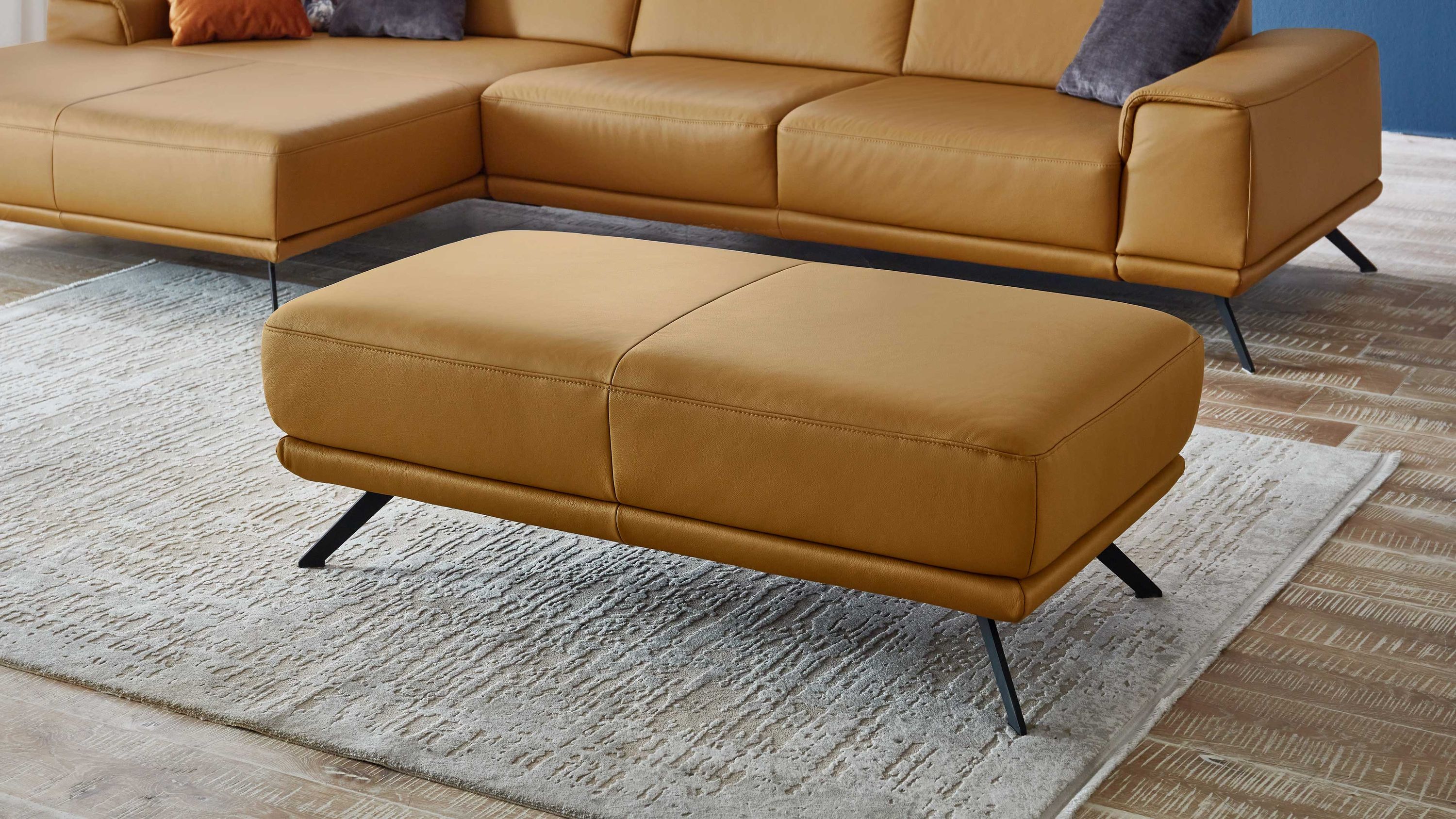 XXL-Hocker mit kurkumafarbenem Leder und schwarzen Metallfüßen Rechteckiger Hocker mit kurkumafarbenem Lederbezug und schwarzen Metallfüßen vor einer passenden Ledercouch