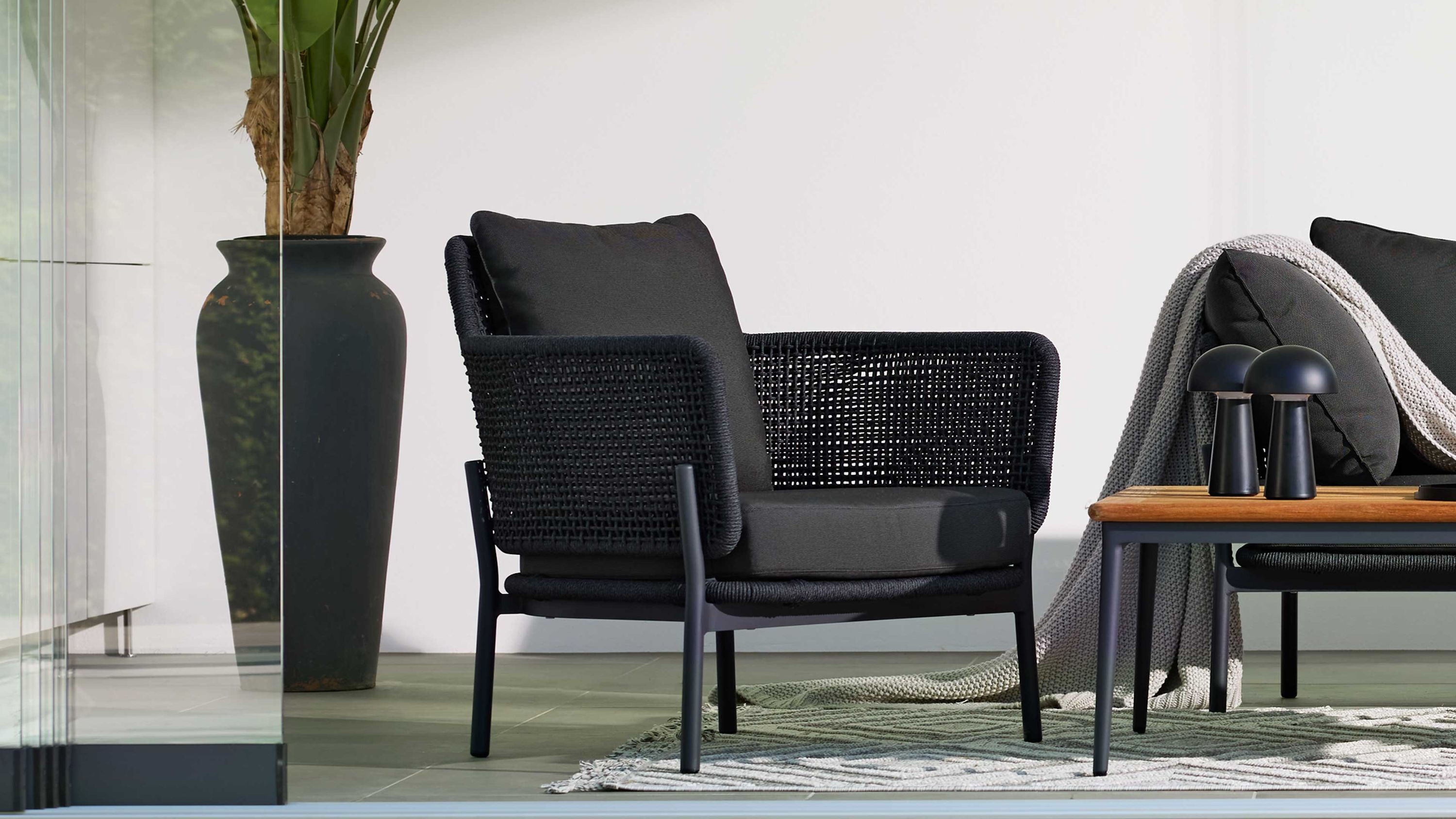 Sessel mit grauem Kordelgeflecht und dunkelgrauem Bezug neben Tisch mit Deko und Sofa mit Kissen und Decke