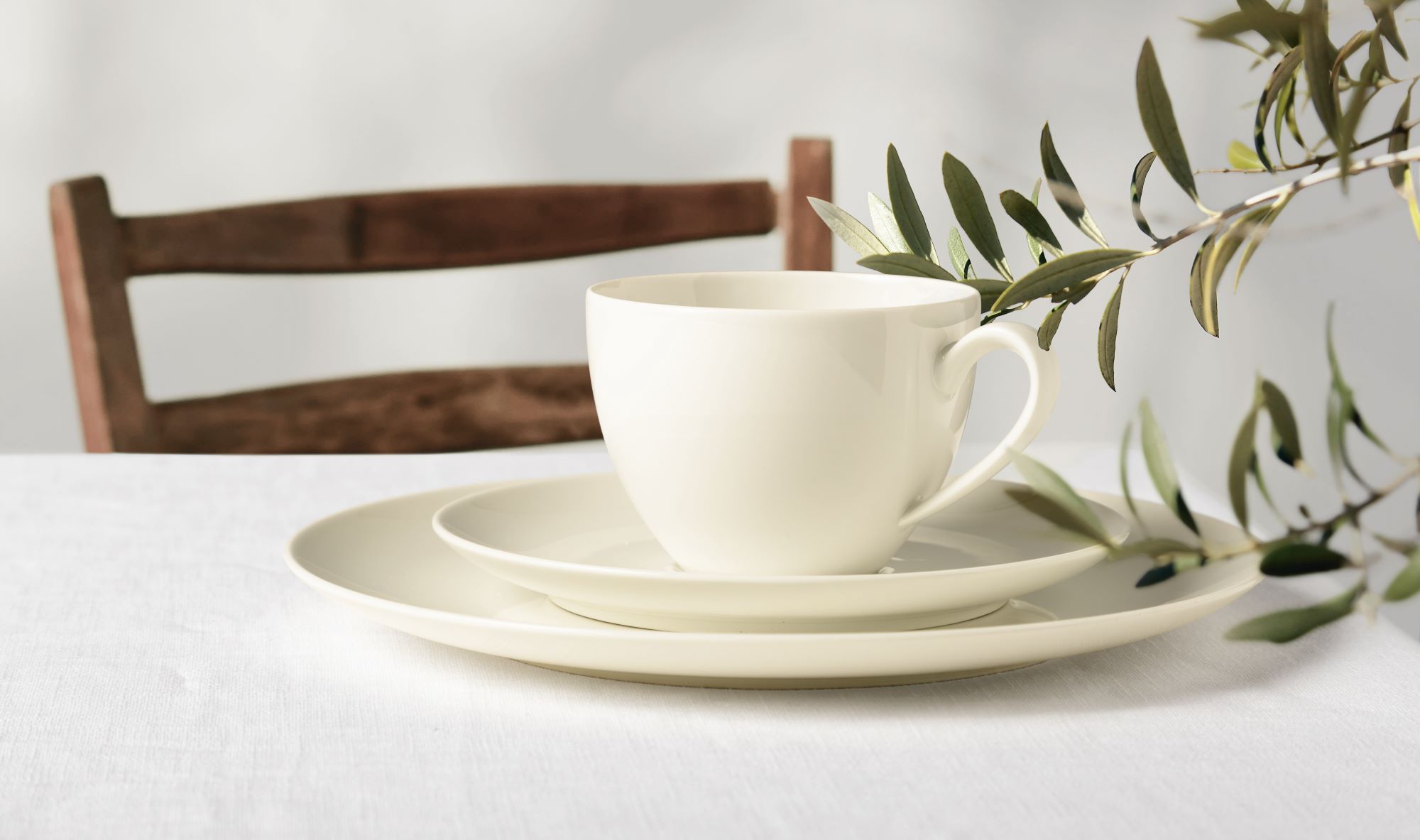 Beige Kaffeetasse mit Henkel auf kleinem und großem Teller auf weißem Tischtuch vor Holzstuhl und Zweig