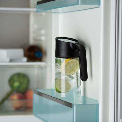 Kühlschrank mit Krug Zitronenwasser im Vordergrund, dahinter Gemüse und weitere Lebensmittel, frontal aufgenommen.