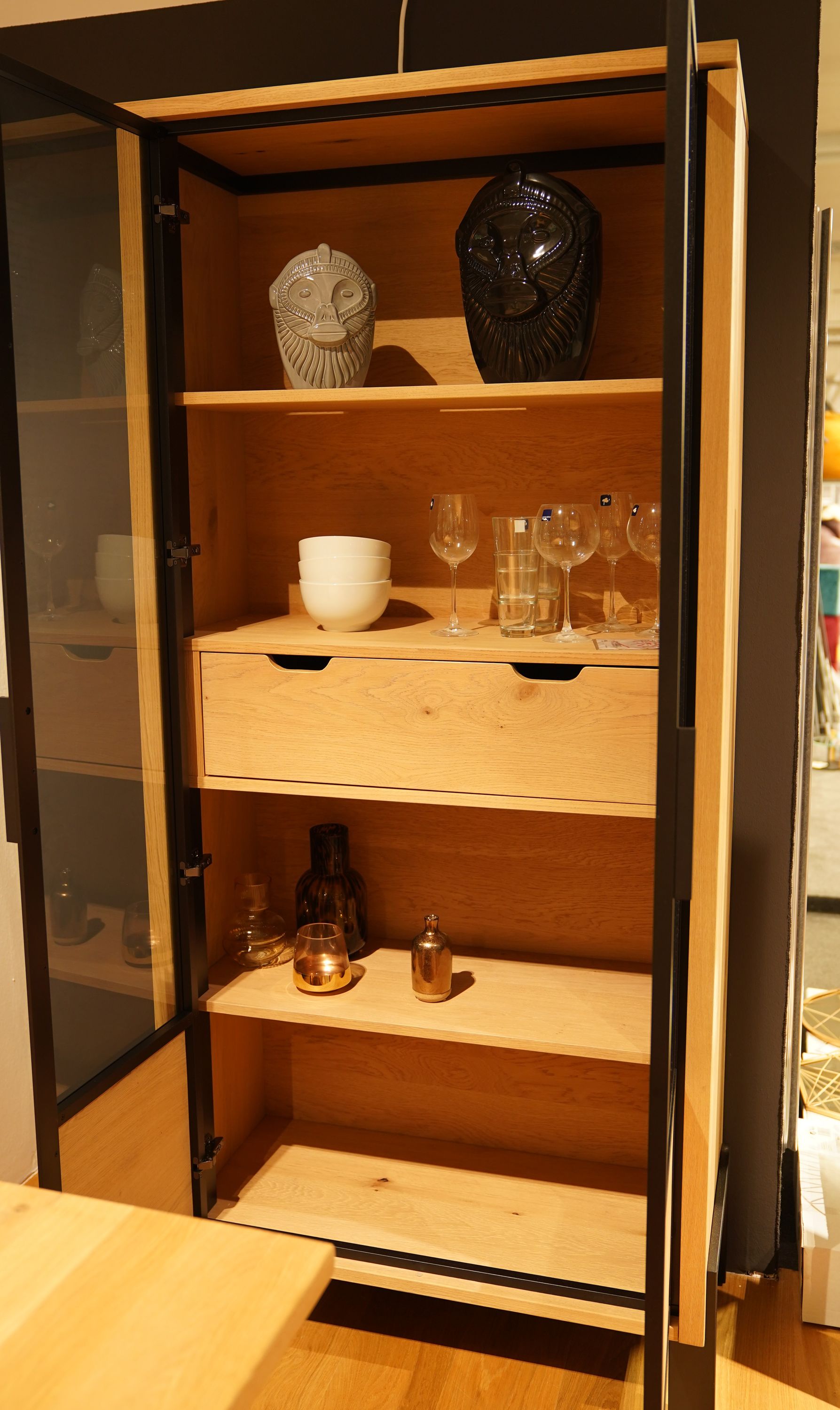 Geöffneter Holzschrank mit Glastüren, befüllt mit Gläsern, Schalen, Vasen und zwei dekorativen Löwenköpfen.