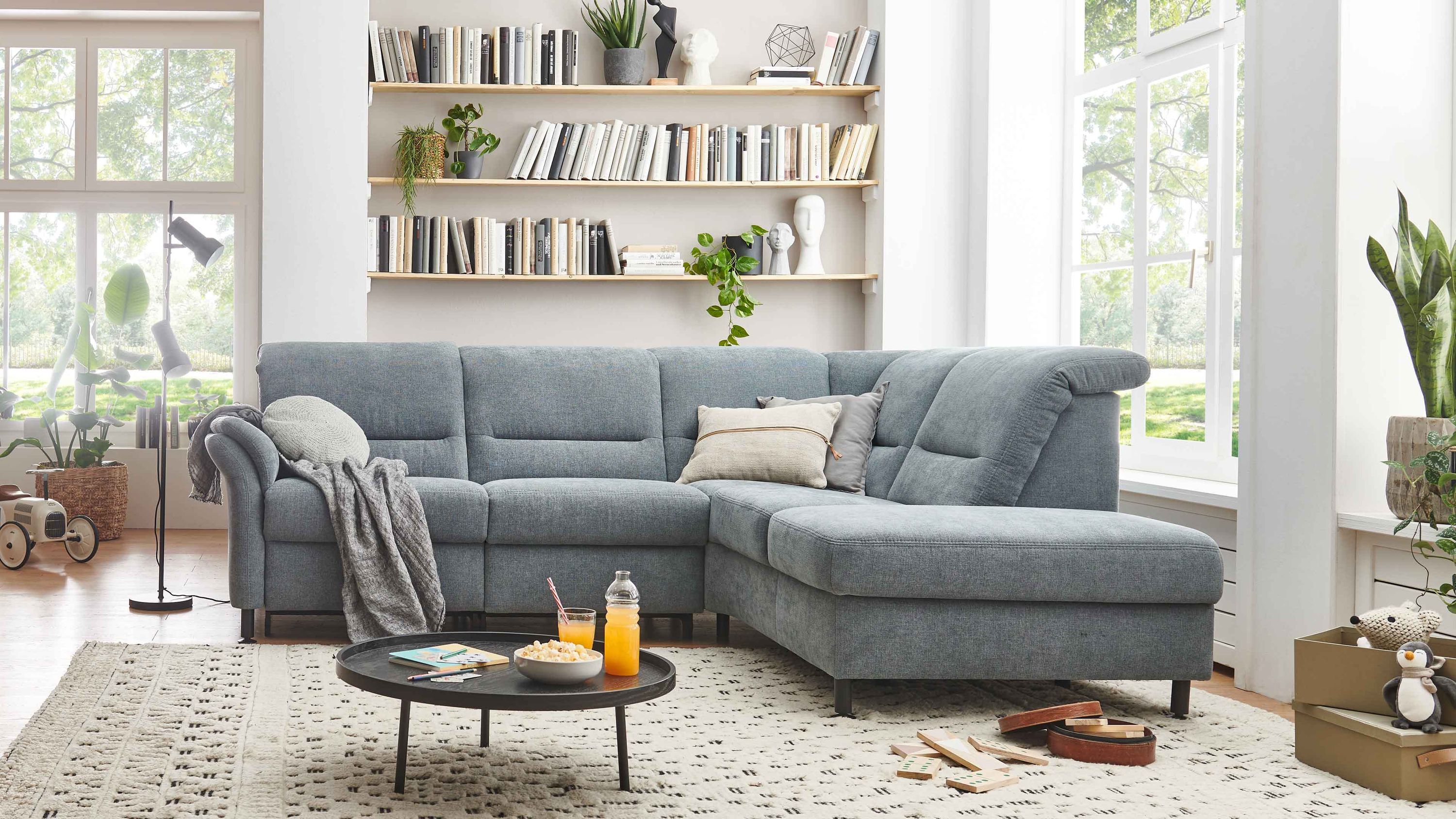 Ecksofa mit stahlfarbenem Stoffbezug und niedrigen Füßen in hellem Wohnzimmer mit Teppich und Bücherregal