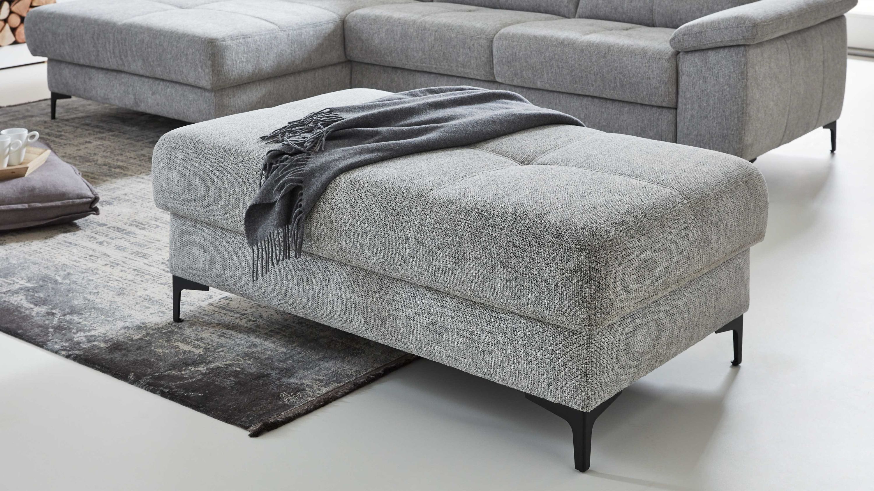 Rechteckiger Polsterhocker mit frostgrauem Flachgewebe und schwarzen Metallfüßen auf Teppich vor grauem Sofa