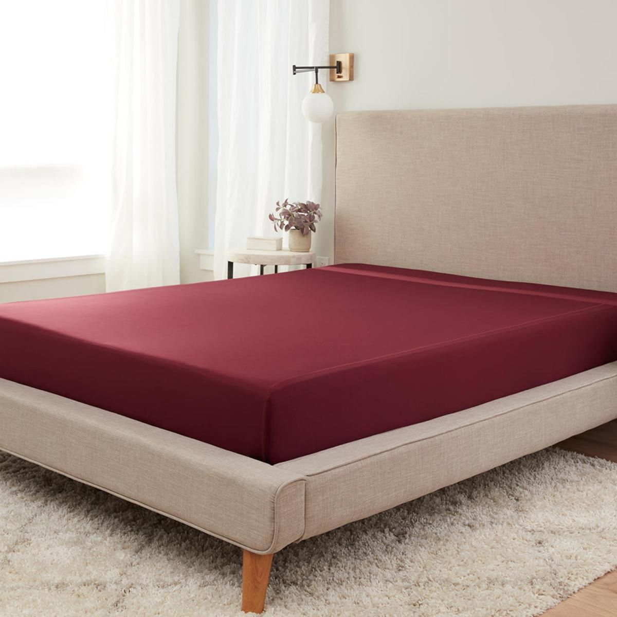 Bett mit Bordeauxfarbenem Spannbetttuch, beige gepolstertes Kopfteil, helle Umgebung
