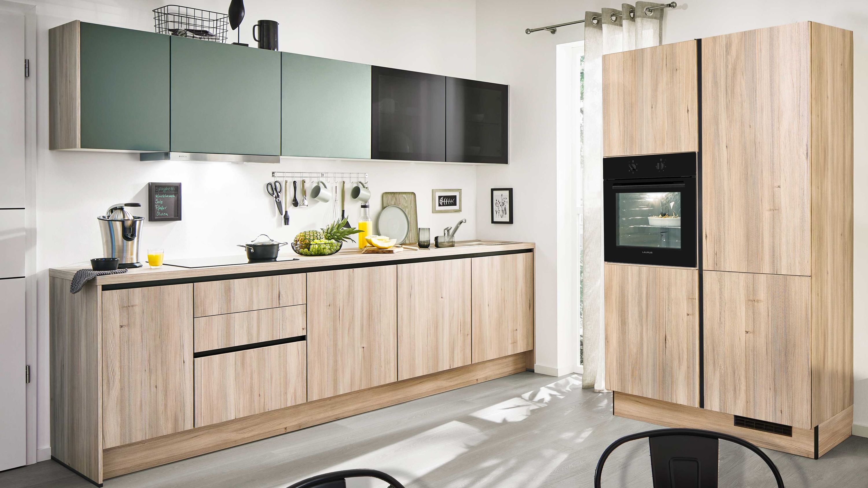 Küche mit Holzfronten und kontrastreichen Oberschränken Helle Holzfronten schwarze und grüne Hängeschränke Einbauofen Fenster mit Vorhang Küchenutensilien Tageslicht.