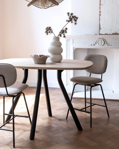 Moderner Esstisch mit Dekovase vor klassischem Kamin. Runder Esstisch mit Holzplatte und schwarzen Beinen, zwei Polsterstühle, Vase mit Trockenblumen, Kamin im Hintergrund.