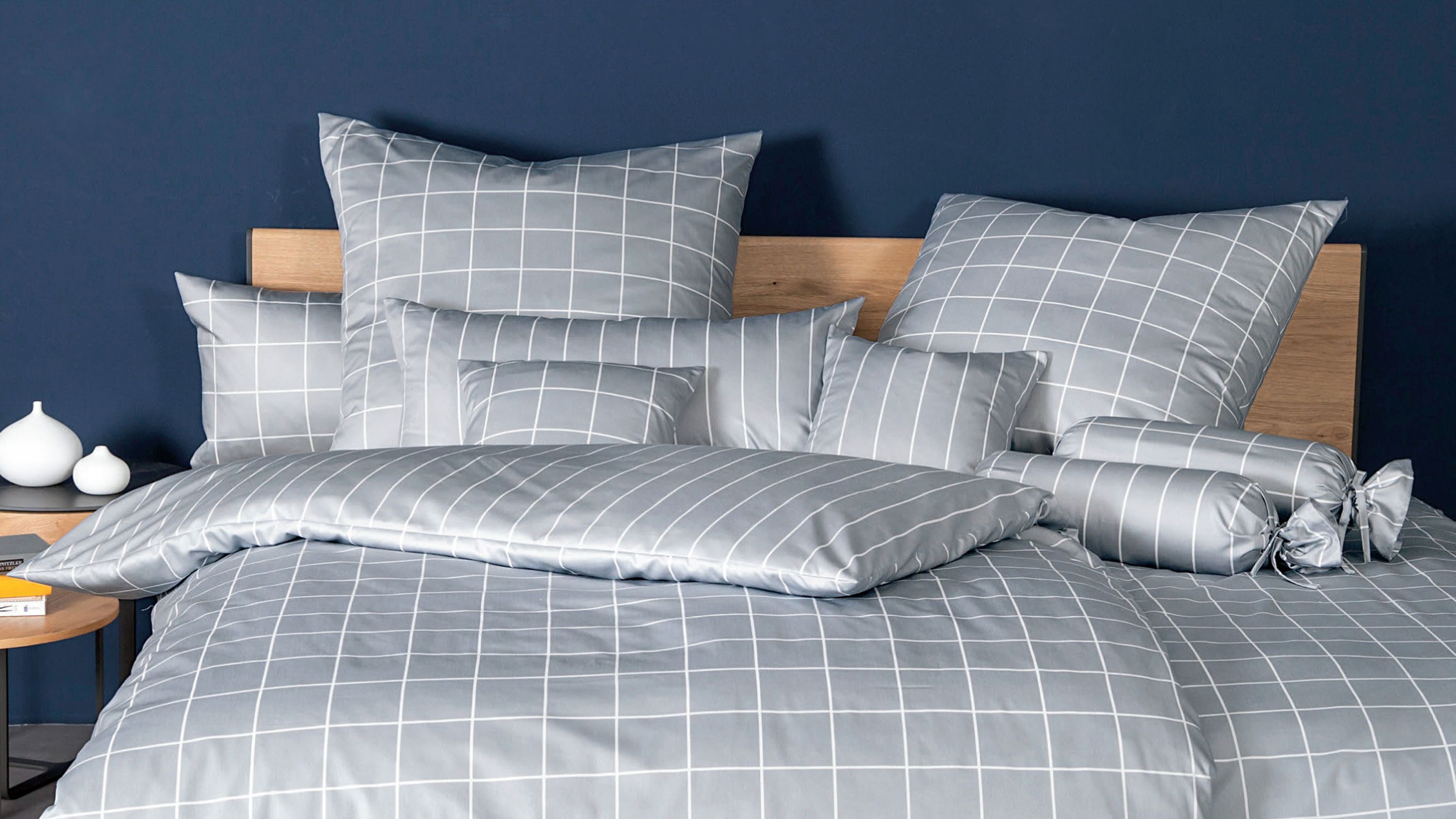 Grau karierte Bettwäsche mit Kissen und Zierrollen auf einem Bett mit Holzrahmen vor dunkelblauer Wand
