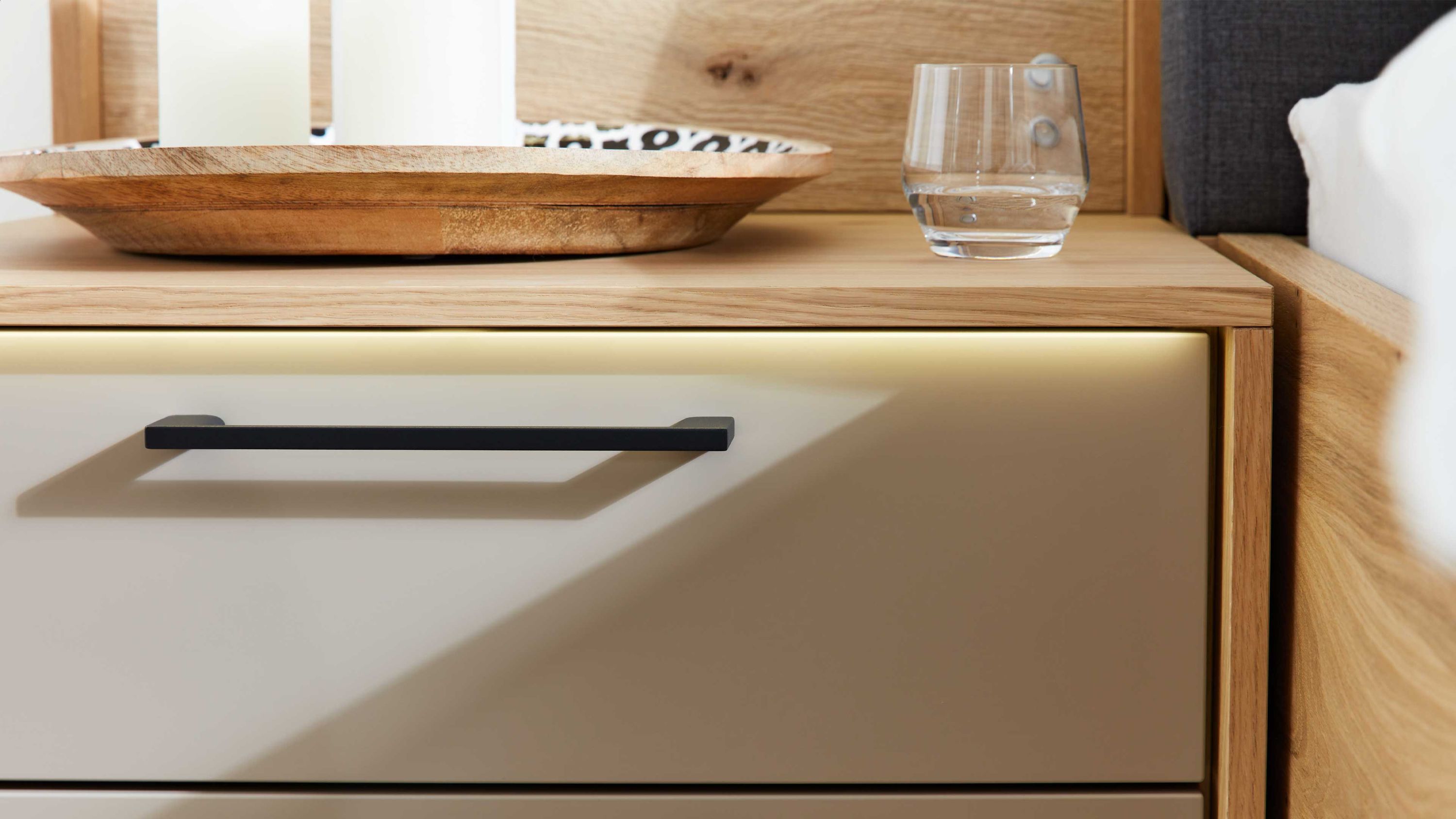 Beige Nachttisch mit schwarzem Griff und integrierter Vorderkantenbeleuchtung neben einem Bett