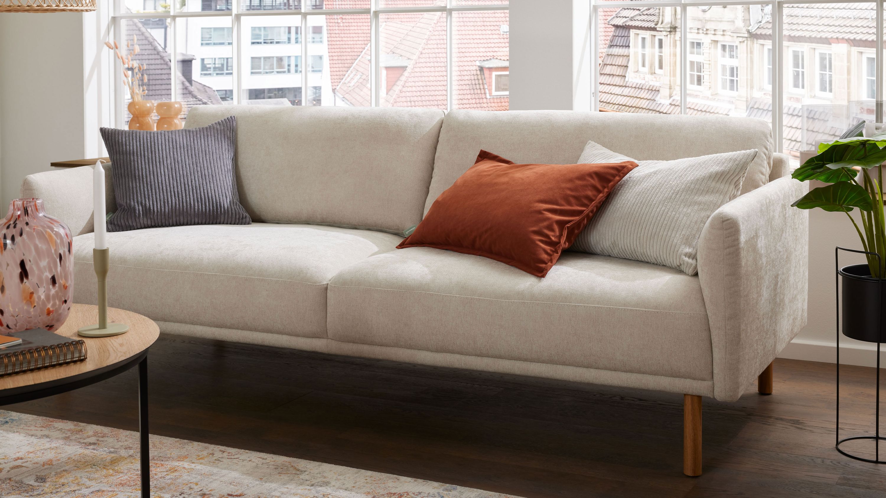Bigsofa mit naturfarbenem Strukturbezug drei Kissen in grau rot und weiß Holzfüße