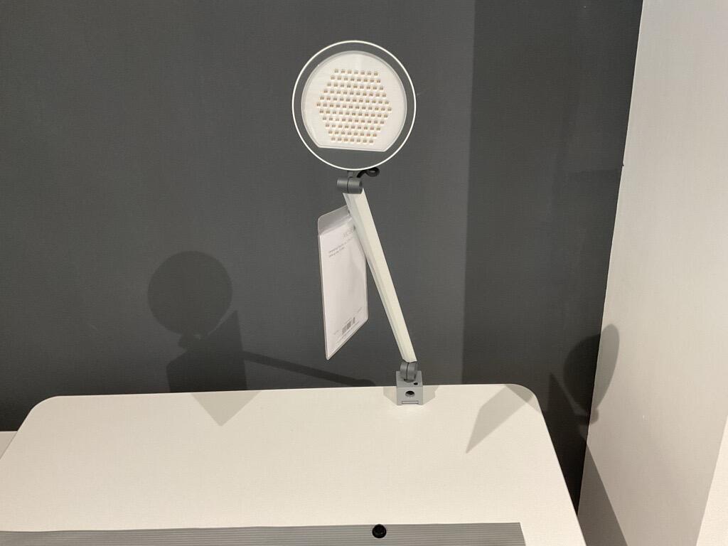 Weiße Tischleuchte mit rundem Waben-Lichtkopf und verstellbarem Arm, frontal an einer Tischkante montiert.