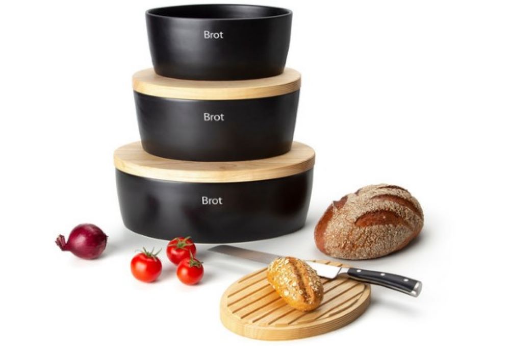 Drei schwarze ovale Brottöpfe mit Holzdeckel und weißer Brot-Aufschrift  Brot und Gemüse auf Holzbrett davor