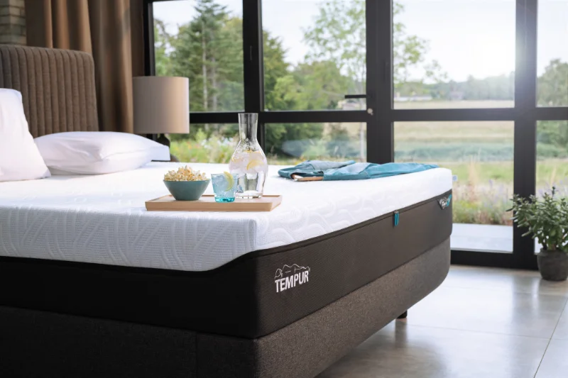 Hochwertiges TEMPUR Boxspringbett in modernem Schlafzimmer mit weißer Matratze, strukturiertem Bettrahmen, Tablett mit Glas und Karaffe, Tagesdecke und bodentiefen Fenstern mit Blick ins Grüne.