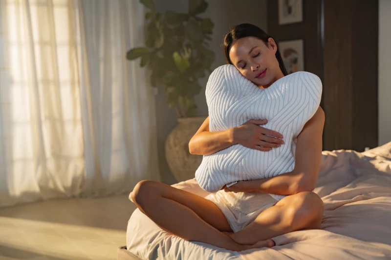 Frau sitzt entspannt im Bett und umarmt ein ergonomisches Tempur Ombracio Schlafkissen im hellen Schlafzimmer.