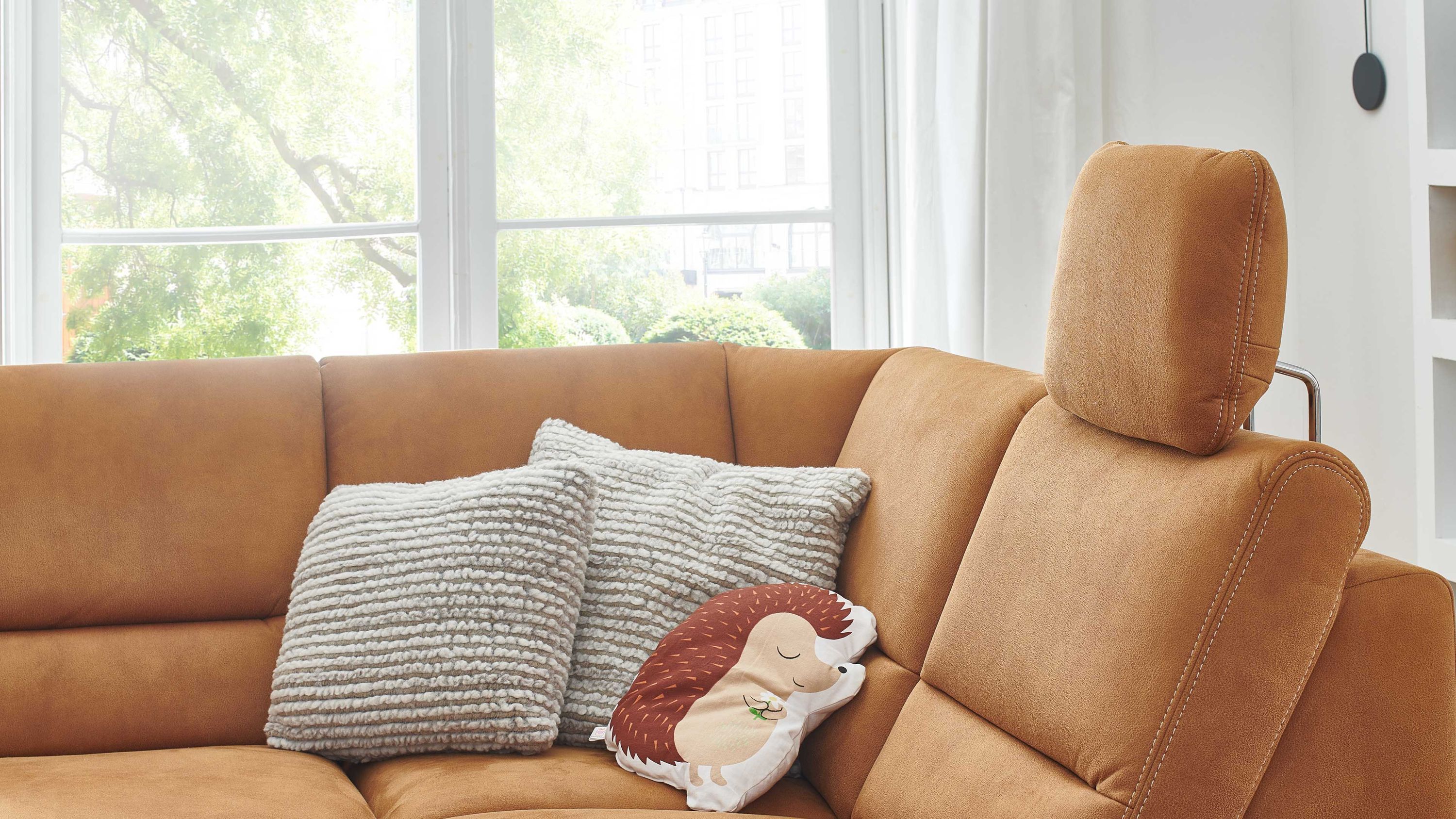 Ecksofa mit Kopfstütze und zimtfarbenem Bezug Ecksofa mit zimtfarbenem Bezug und verstellbarer Kopfstütze zwei graue Kissen und ein Igel-Kissen